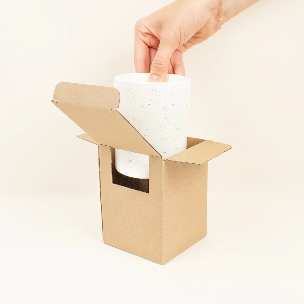 Taza de cerámica CANDELA Mano sacando una taza de cerámica blanca de una caja de cartón