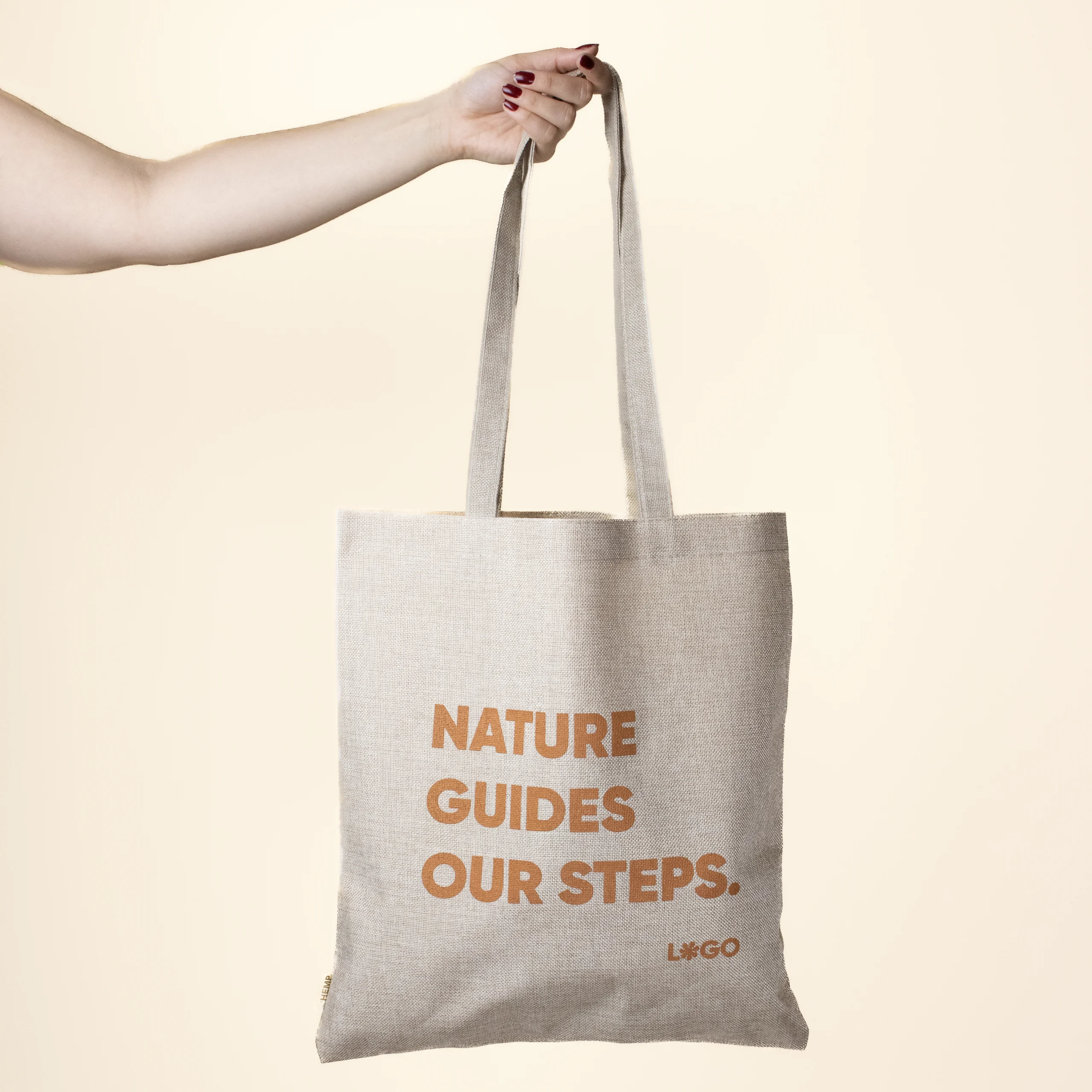 Mano sobre fondo beige sujetando una bolsa de cáñamo personalizada con logo naranja
