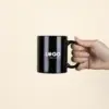 Taza negra personalizada IRIS Mano sujetando una taza negra con asa personalizada con la palabra logo en blanco fotografiada sobre un fondo beige