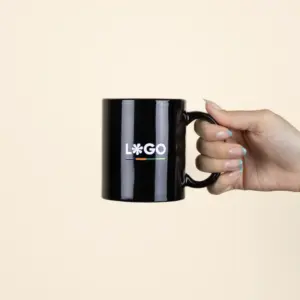 Mano sujetando una taza negra con asa personalizada con la palabra logo en blanco fotografiada sobre un fondo beige