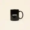 Taza negra personalizada IRIS Taza negra con asa personalizada con la palabra logo en blanco fotografiada sobre un fondo beige