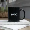 Taza negra personalizada IRIS Taza negra con asa personalizada con la palabra logo en blanco colocada encima de un cuaderno blanco