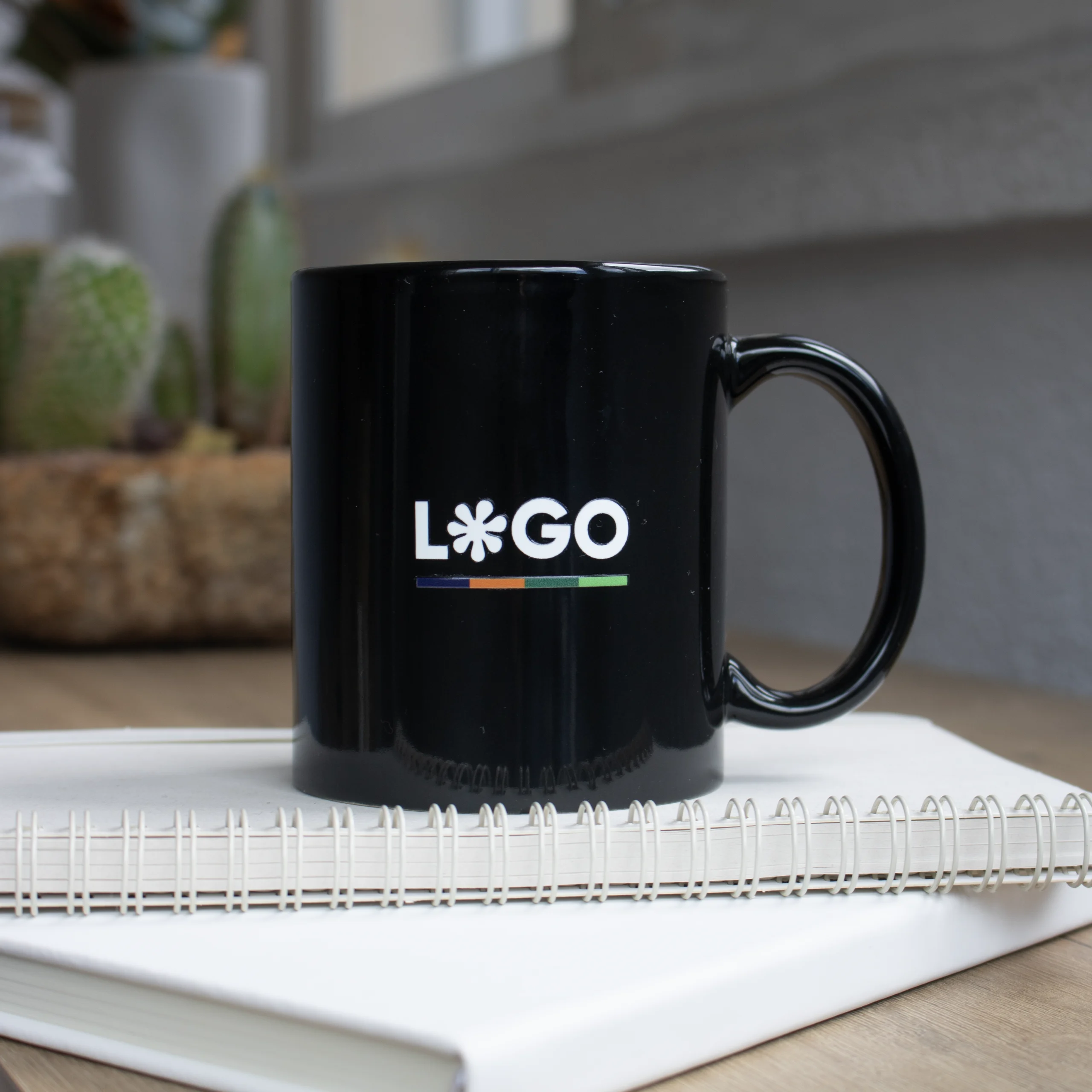 Taza negra personalizada IRIS Taza negra con asa personalizada con la palabra logo en blanco colocada encima de un cuaderno blanco