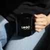 Taza negra personalizada IRIS Chica en vaqueros agarrando una taza negra con asa personalizada con la palabra logo en blanco