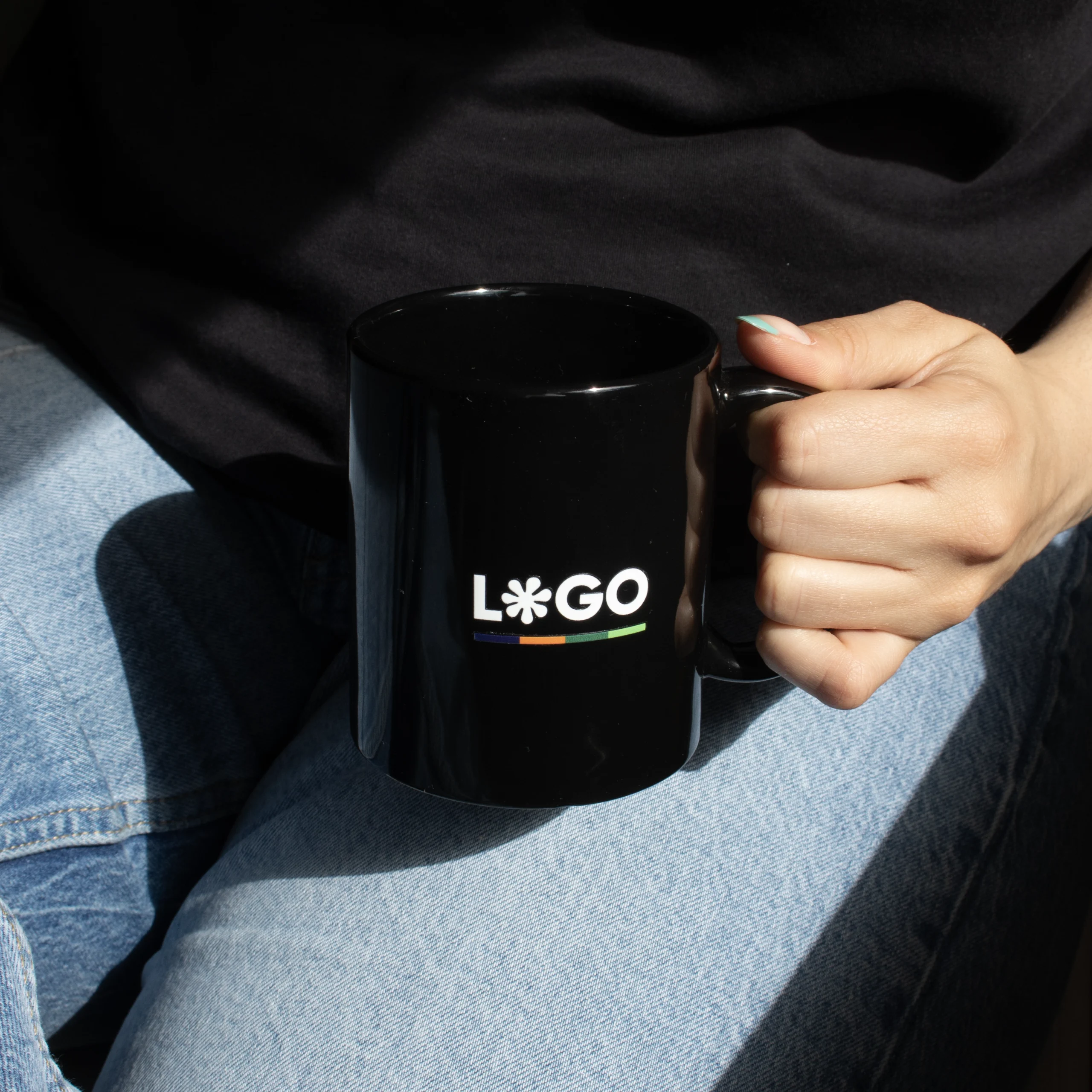 Taza negra personalizada IRIS Chica en vaqueros agarrando una taza negra con asa personalizada con la palabra logo en blanco