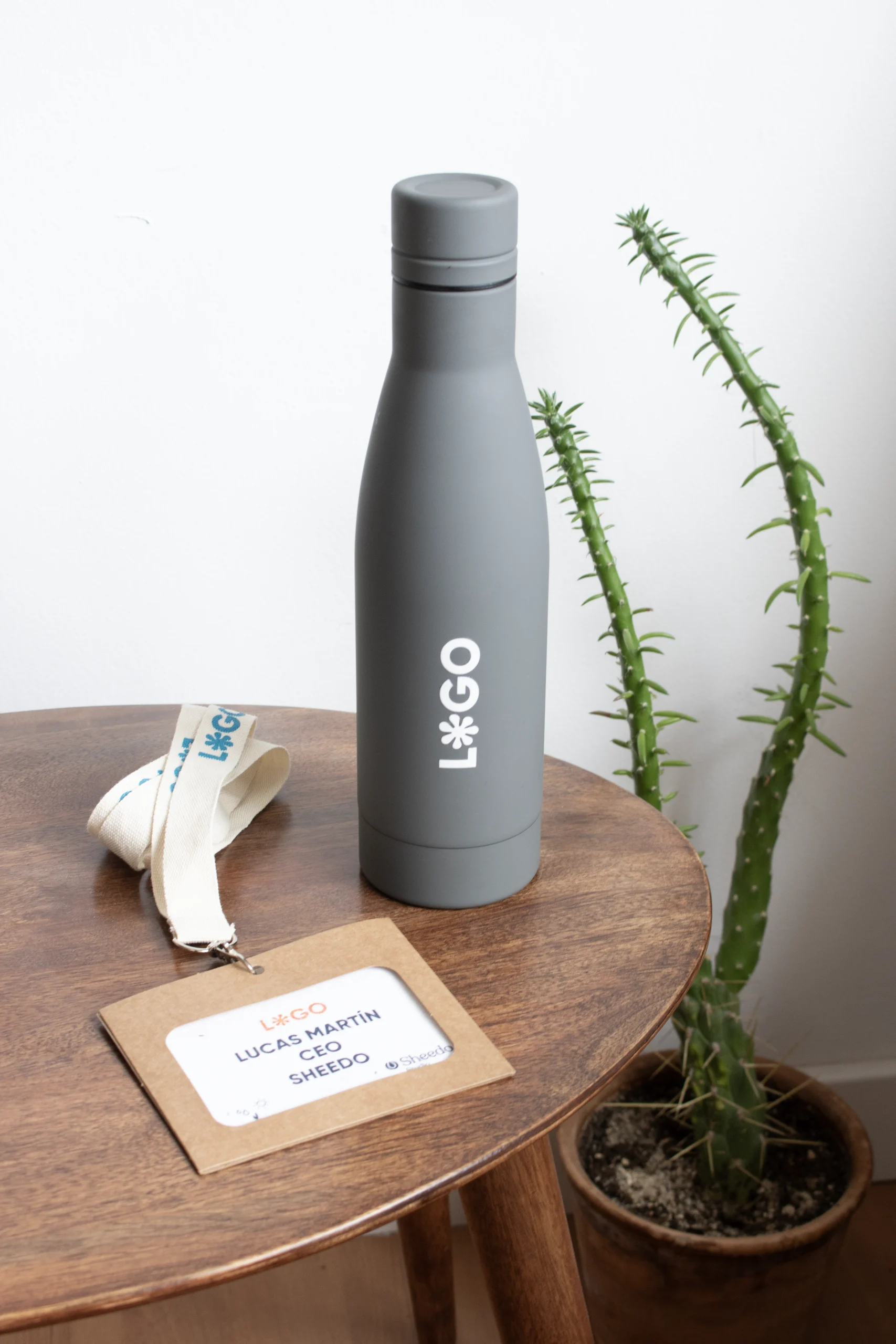 Botella gris de acero inoxidable personalizada con logo blanco junto a una acreditación con lanyard sobre una mesa de madera
