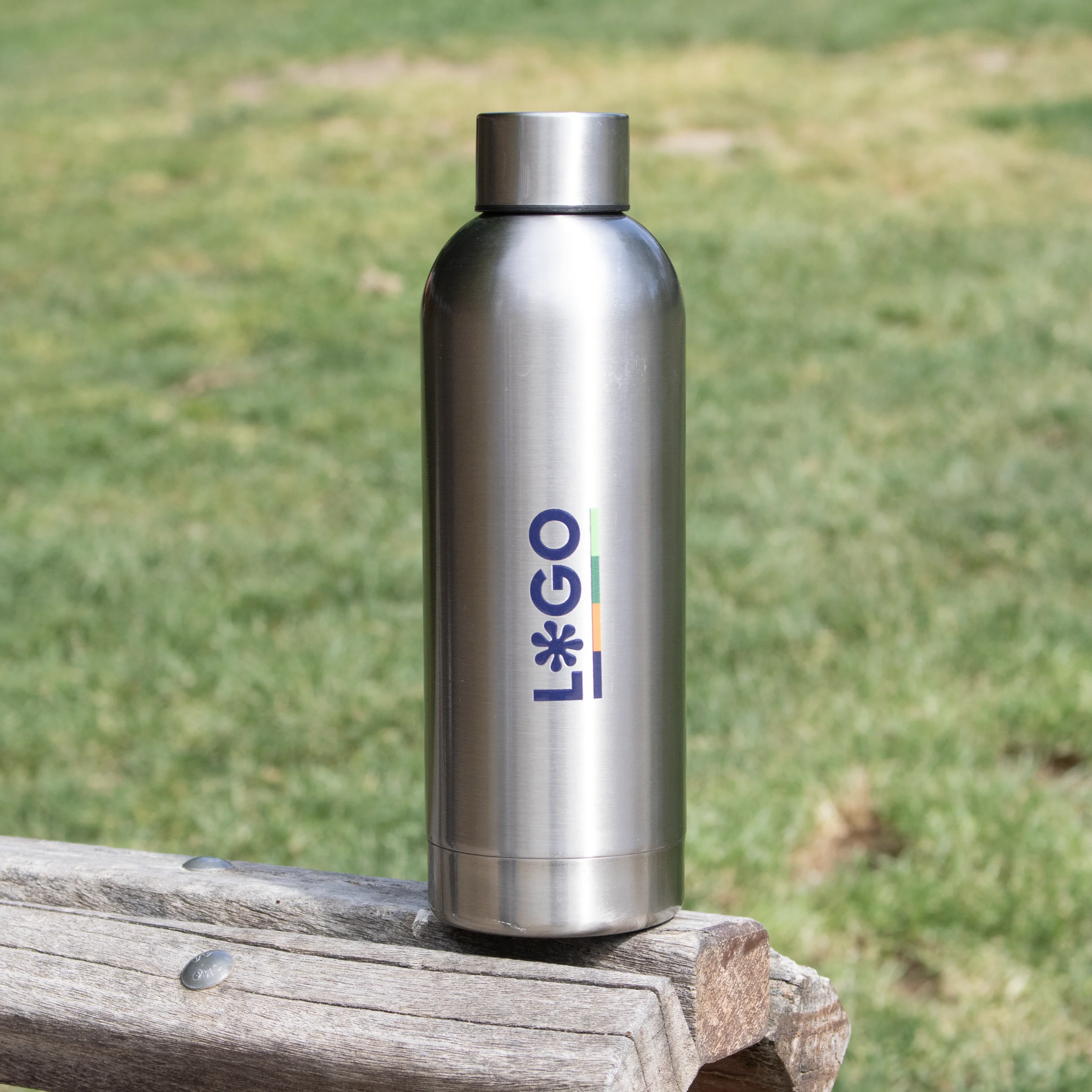 Botella acero inoxidable personalizada con logo sobre banco de madera fondo parque hierba
