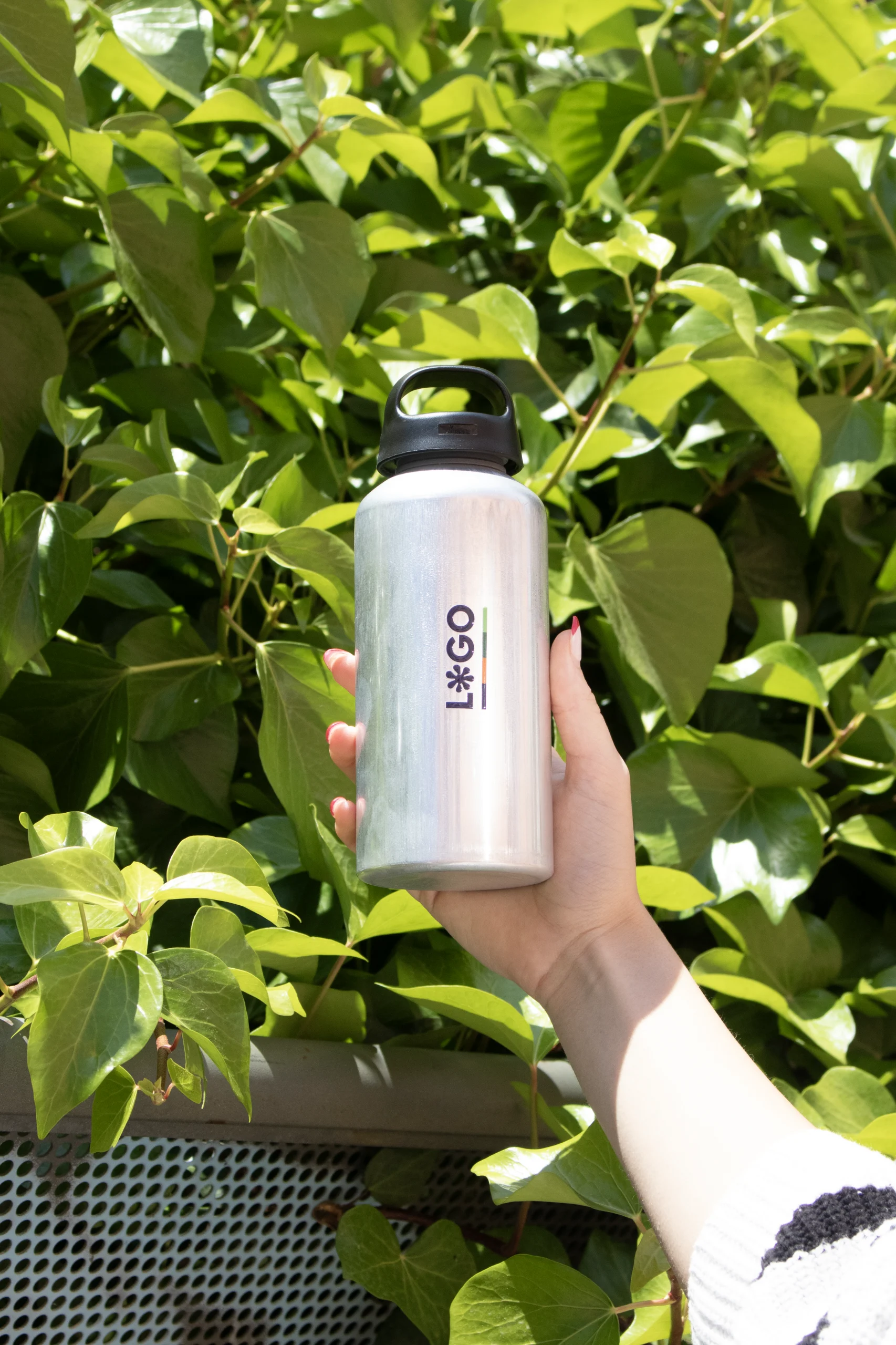 Mano sujetando una botella de aluminio personalizada con la palabra logo con fondo de plantas