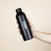 Botella térmica color negro NORMA EN USO Botella térmica color negro personalizada con logo blanco modelo mano fondo beige