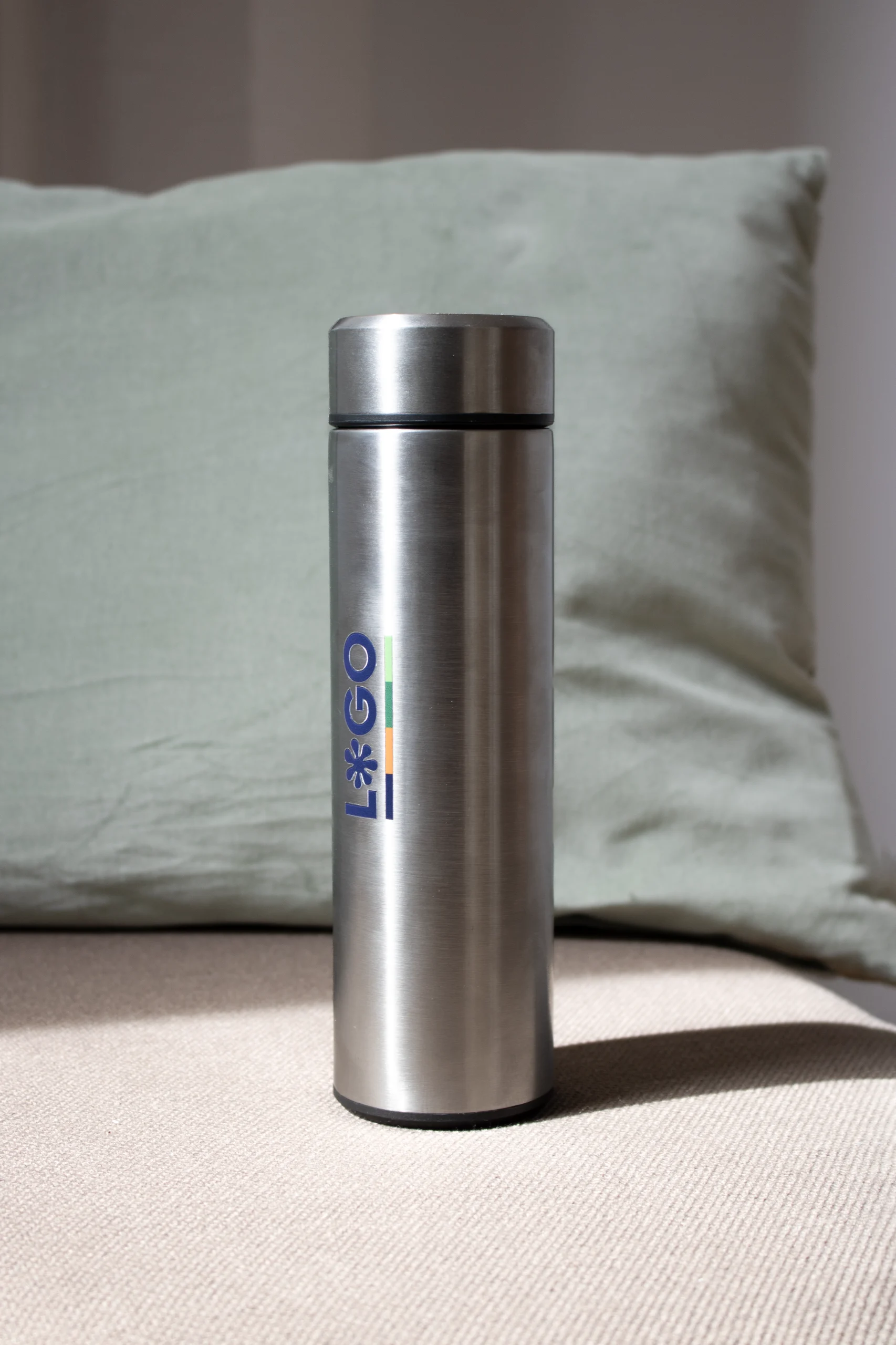 Termo acero inoxidable personalizado plateado logo azul delante de cojín verde
