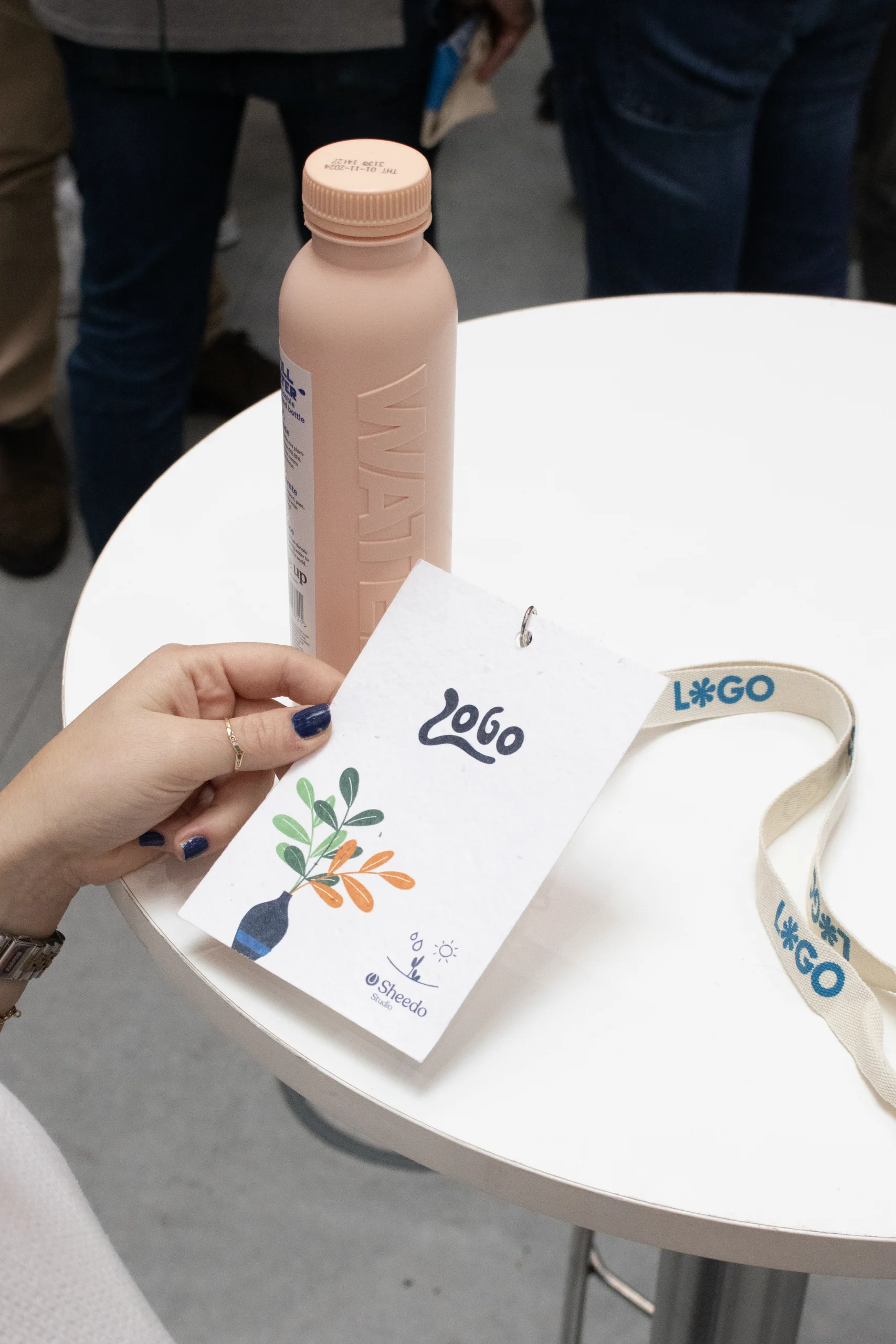 Mano sujetando una acreditación con lanyard y una botella de agua rosa en un evento de empresa