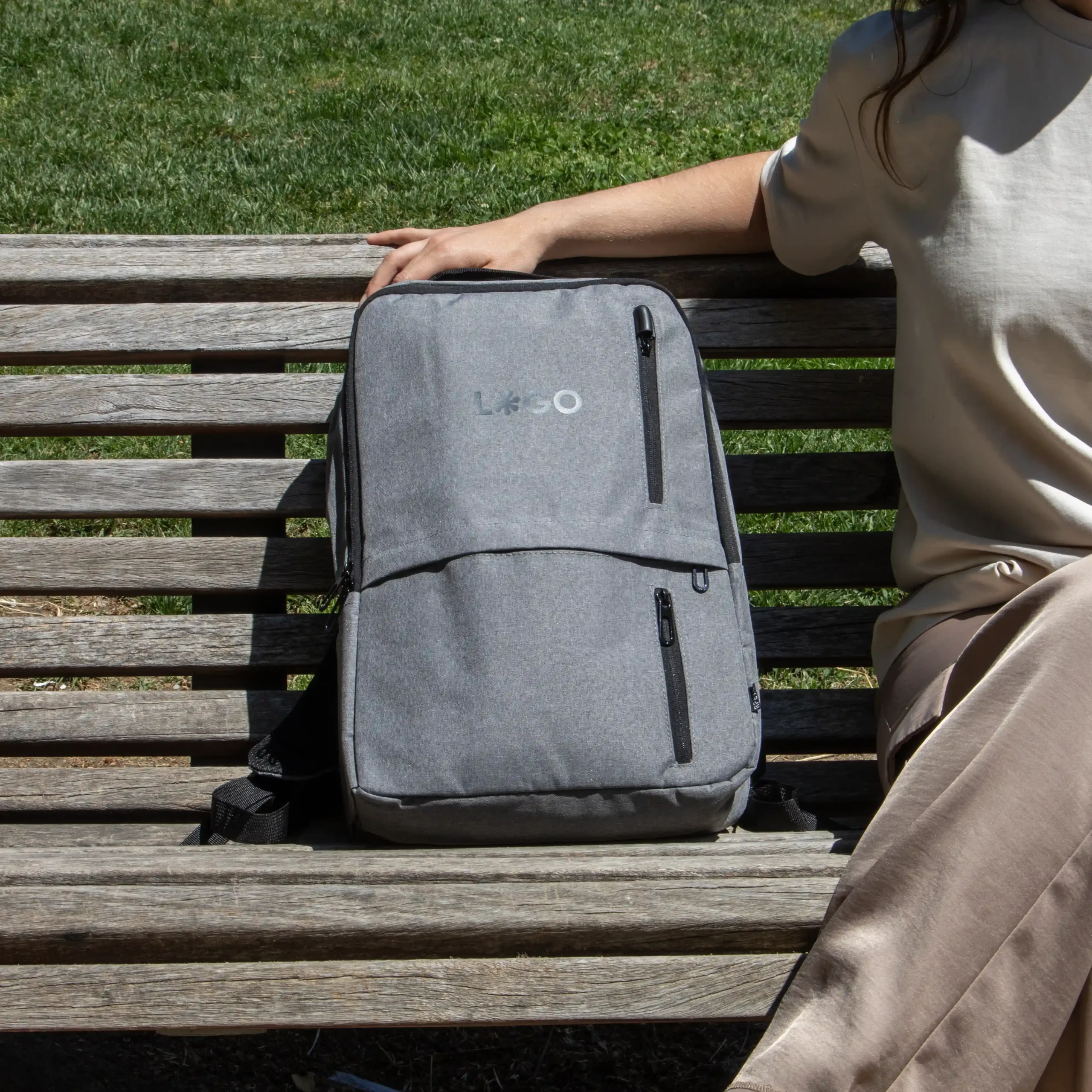 Mochila de empresa DUPPIA Gris Sol Mochila de empresa personalizada gris con logo negro modelo chica sentada en banco parque