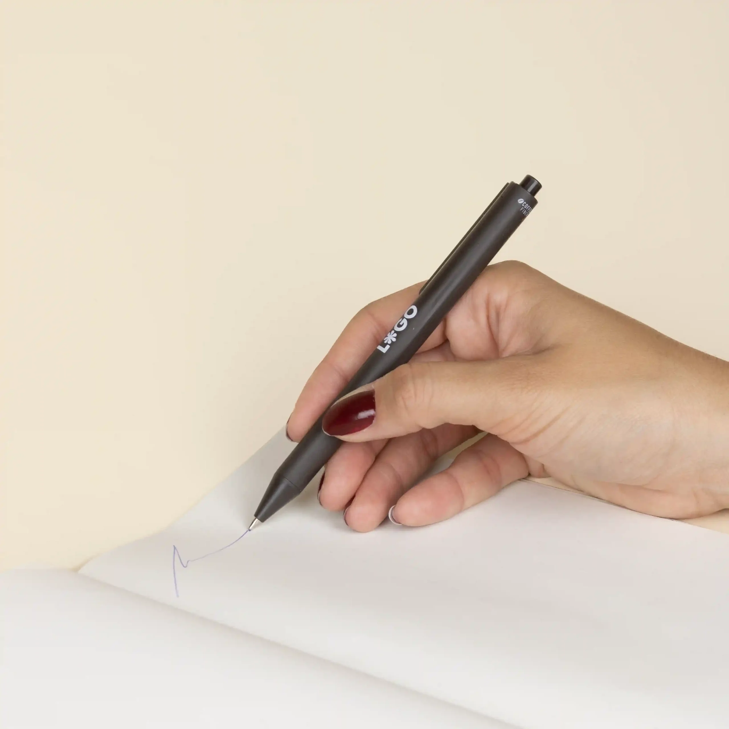 Mano escribiendo en cuaderno con bolígrafo de fibra de cafe personalizado con logo blanco