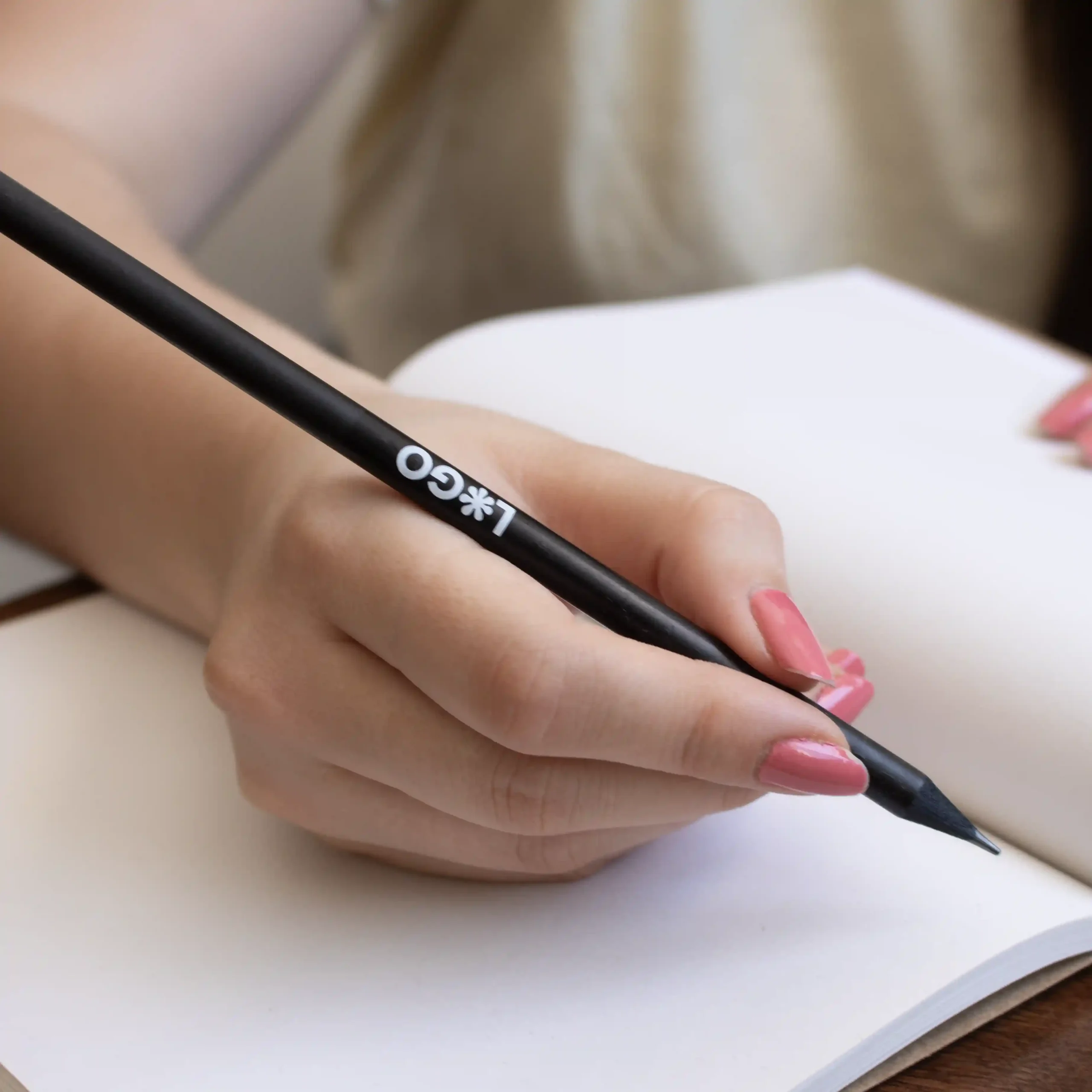 Mano escribiendo en cuaderno con lápiz de madera negro personalizado con logo blanco