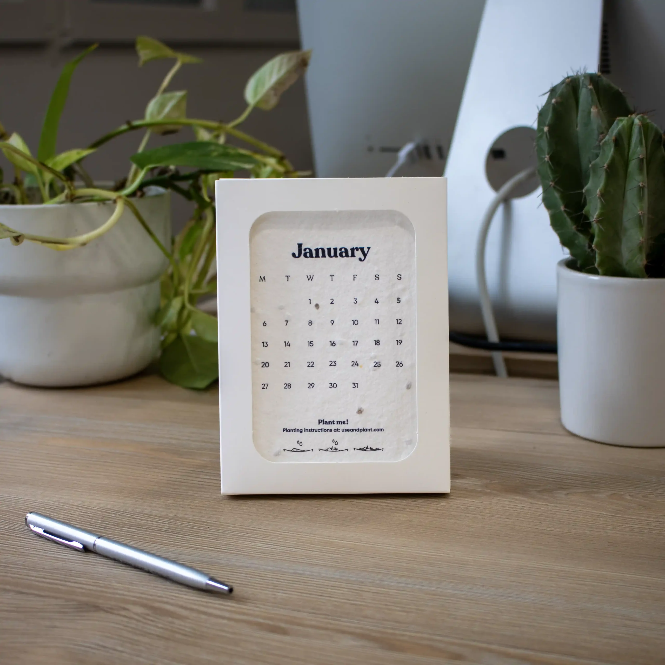 Calendario de semillas en estuche blanco personalizado colocado en una mesa de trabajo con un bolígrafo y un ordenador