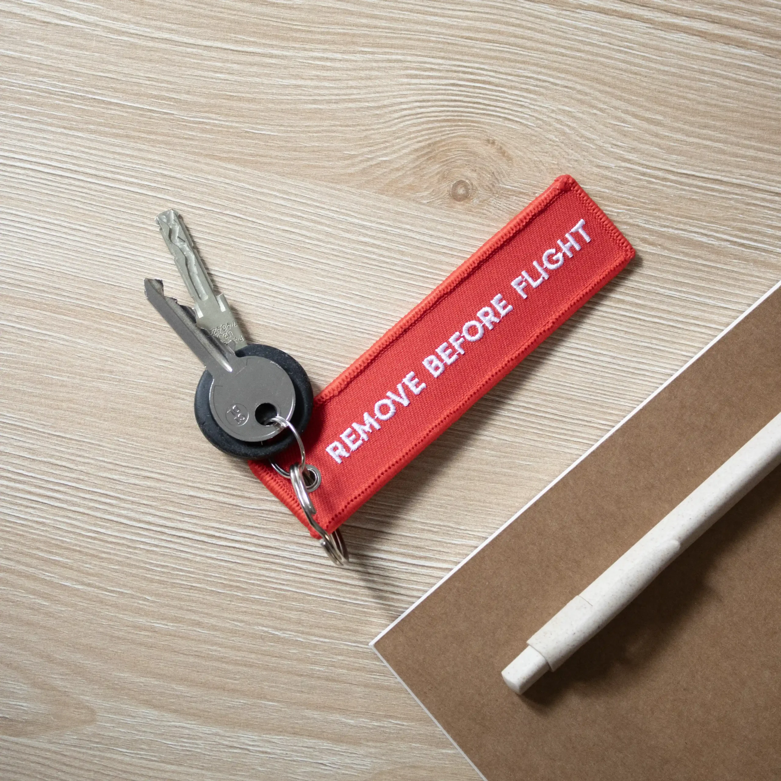 Llaves con llavero de tela rojo bordado con hilo blanco sobre una mesa de madera junto a un cuaderno y un bolígrafo