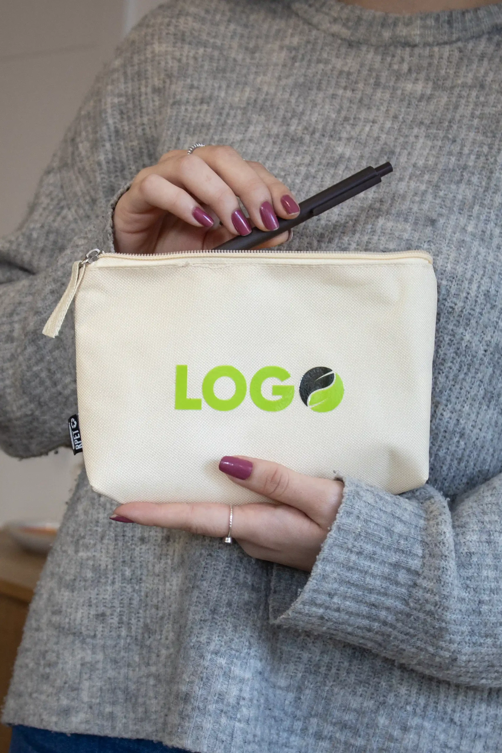 Chica con jersey gris mostrando un neceser personalizado con logo verde y un bolígrafo marrón