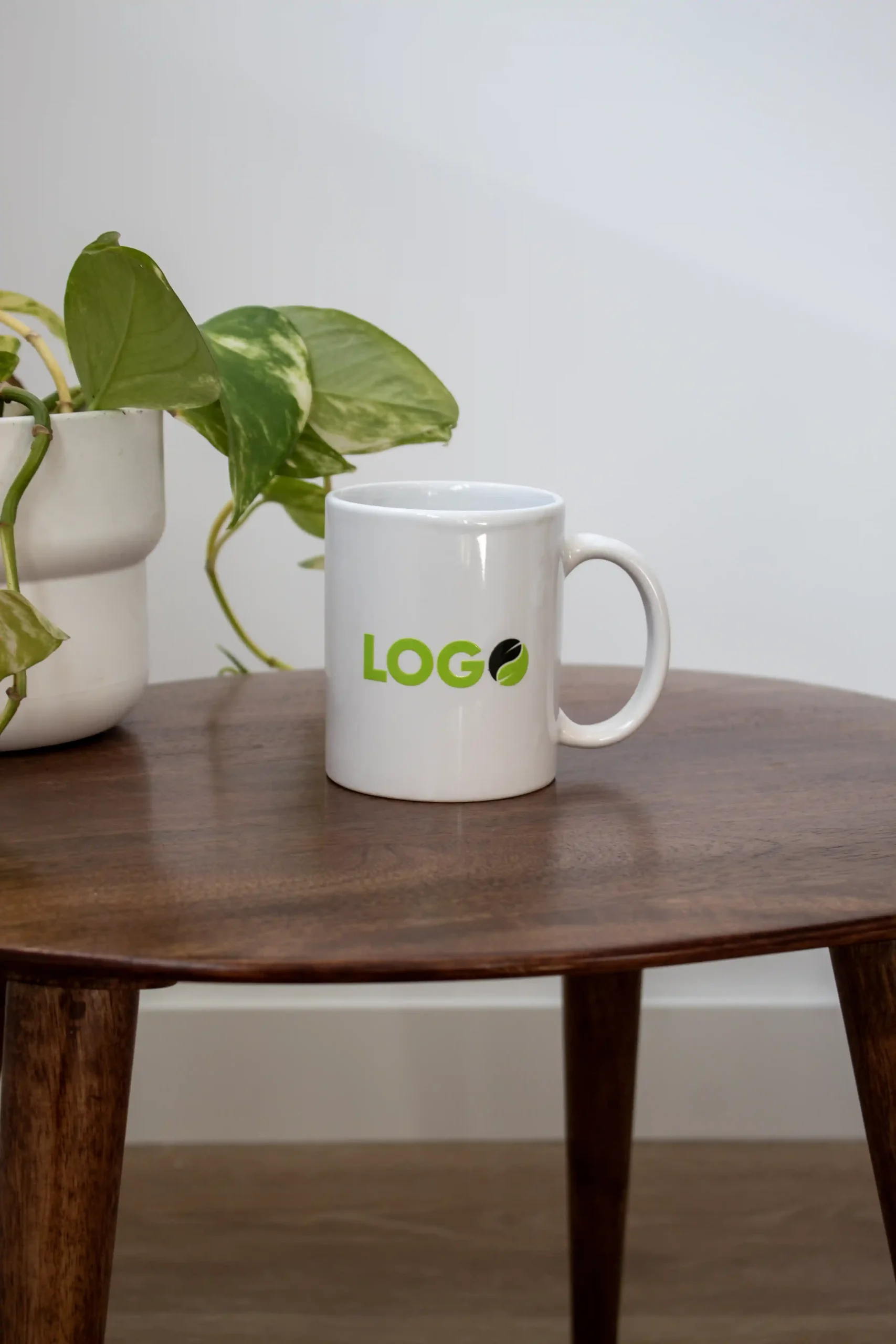 Taza blanca personalizada con la palabra logo en verde junto a una planta apoyadas en una mesa de madera