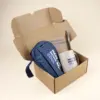 Caja de cartón abierta con un estuche azul, una taza beige, un cuaderno kraft y un bolígrafo marrón personalizados con logo