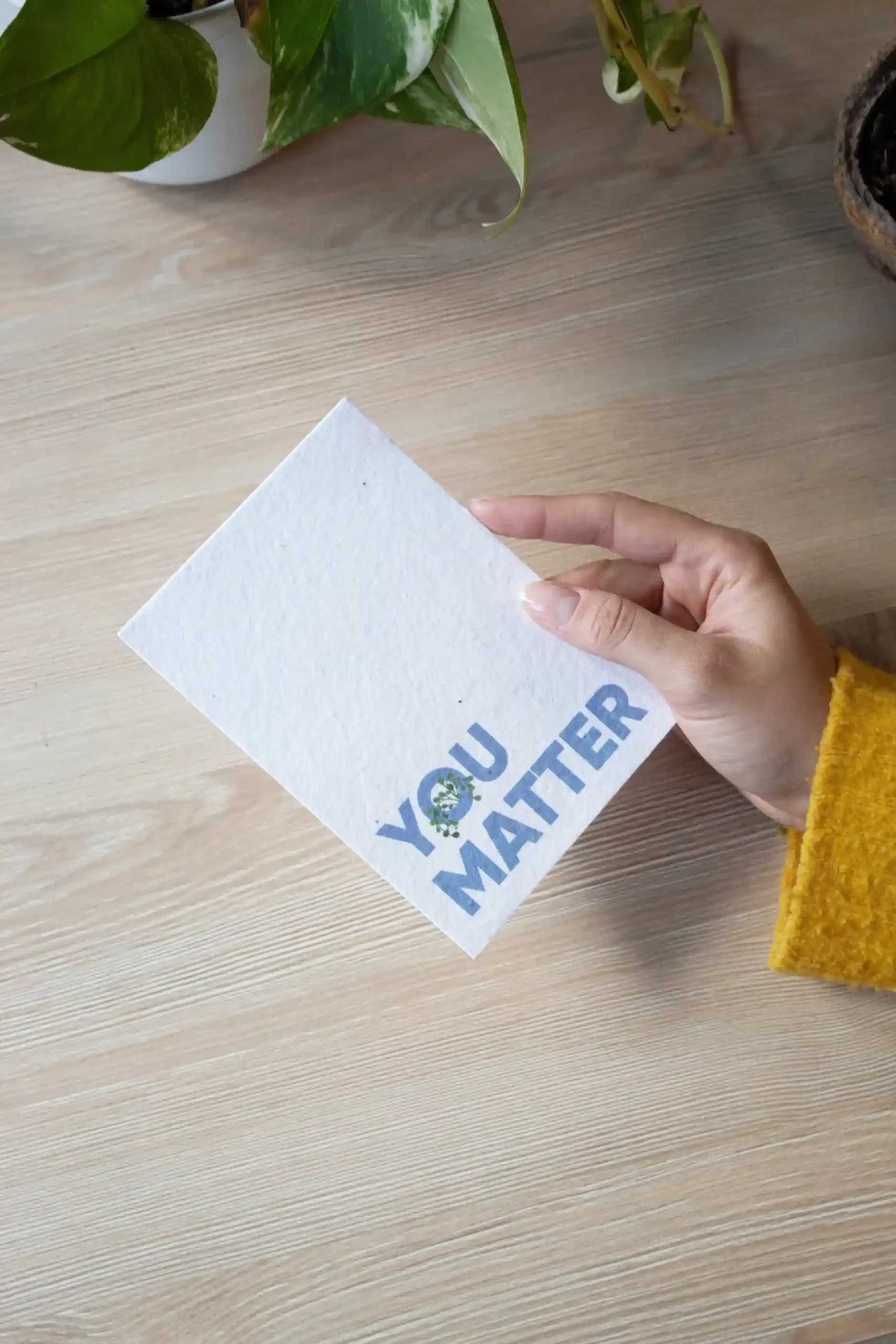 Mano sujetando un flyer A6 de papel plantable blanco personalizado con fondo de mesa de madera