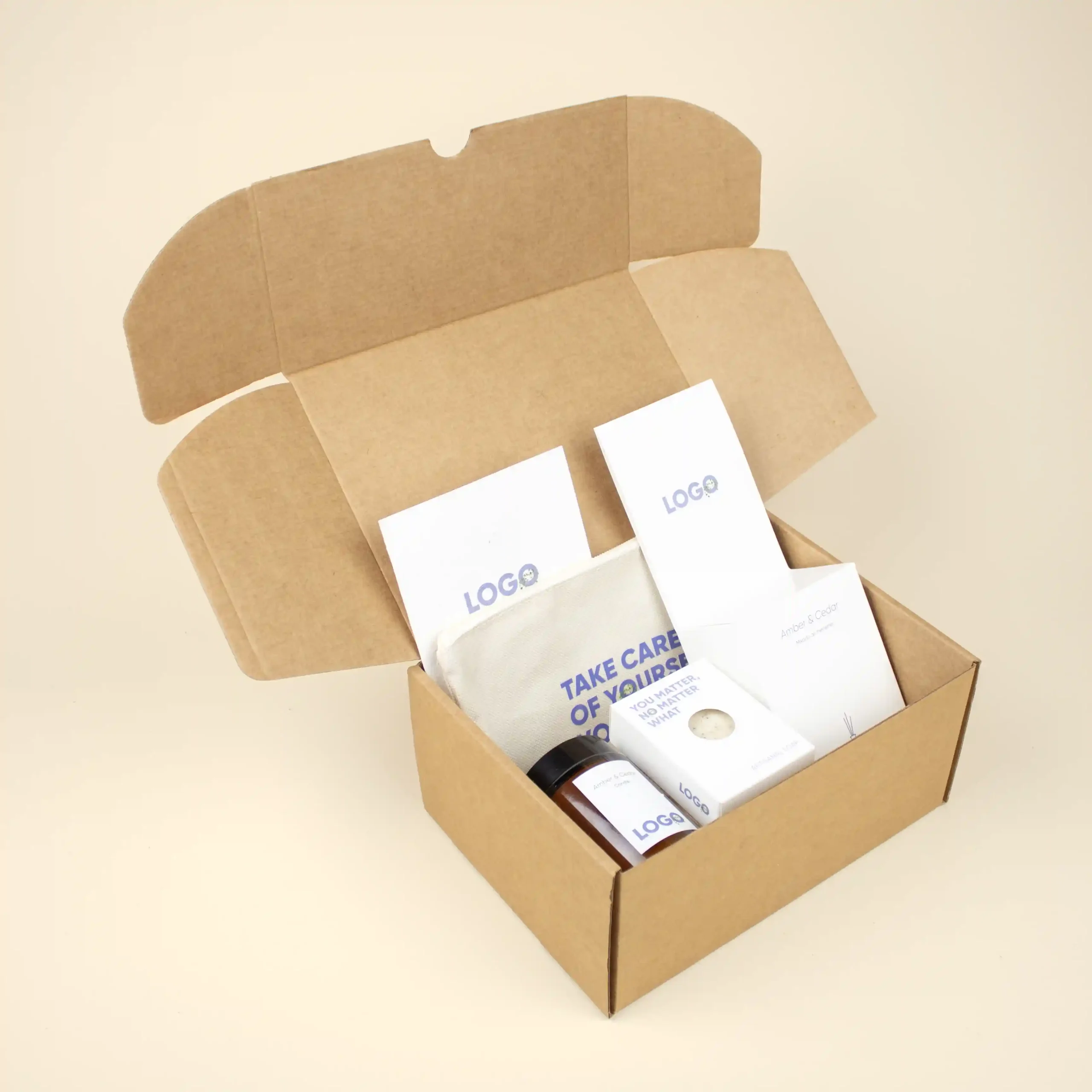 Caja de cartón abierta con un neceser blanco, un ambientador mikado, una vela en tarro de cristal, un jabón ecológico y un flyer A6 personalizados