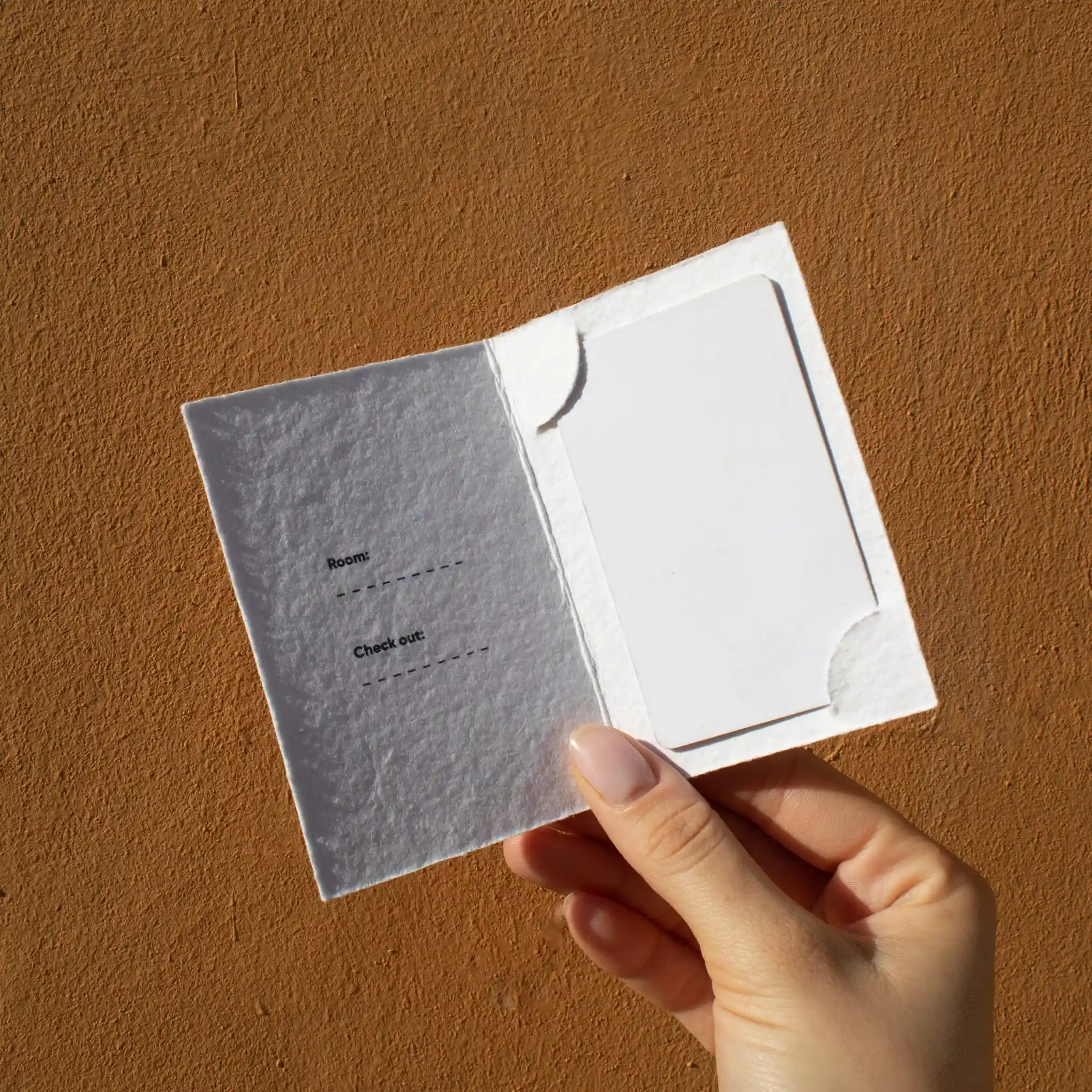 Porta llaves hotel díptico KARTE Cotton Mano mostrando un porta llaves de hotel díptico en papel ecológico blanco abierto con una tarjeta blanca dentro