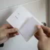 Porta llaves hotel díptico KARTE Cotton Mano delante de una puerta de hotel mostrando un porta llaves de hotel en papel ecológico blanco abierto con una tarjeta blanca dentro