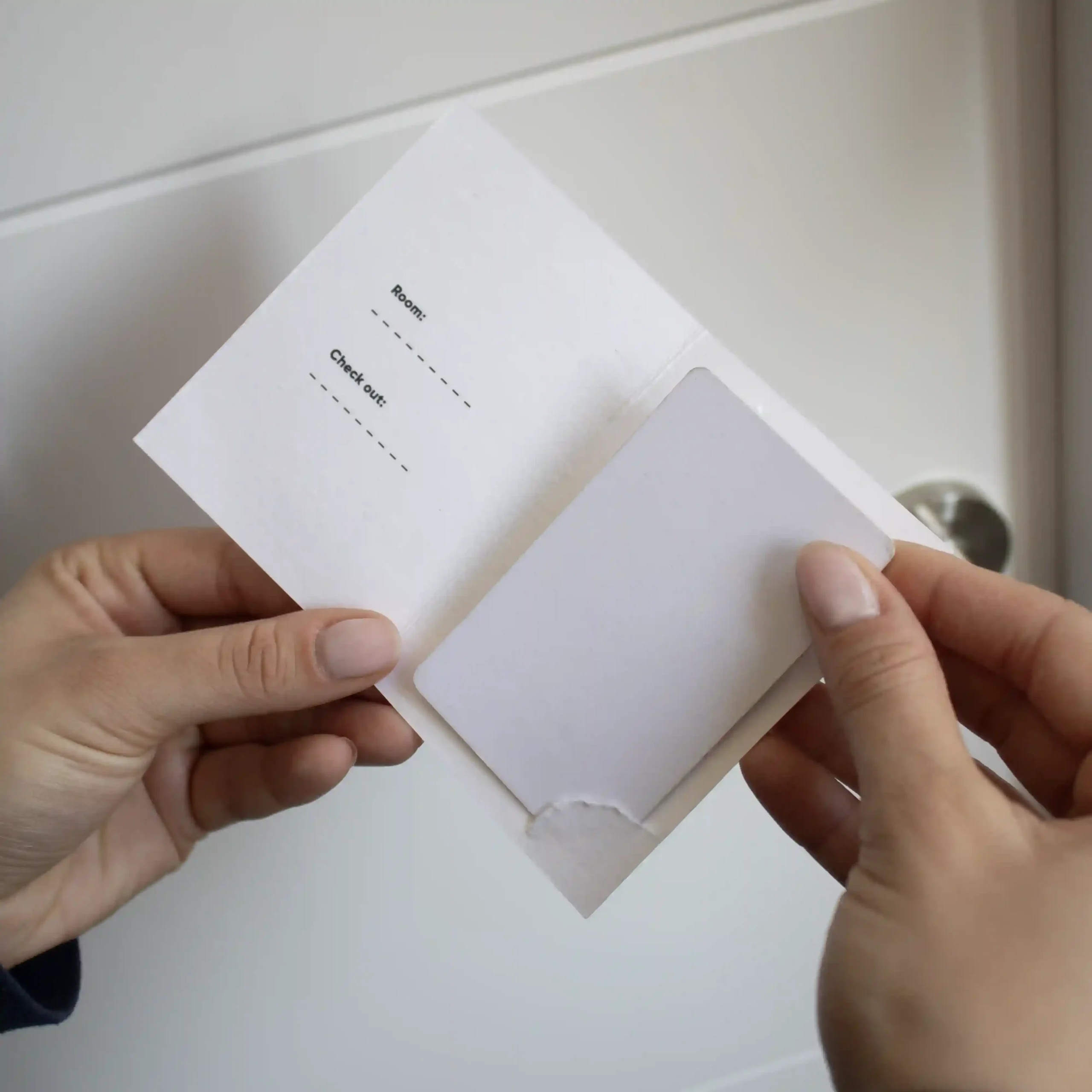 Porta llaves hotel díptico KARTE Cotton Mano delante de una puerta de hotel mostrando un porta llaves de hotel en papel ecológico blanco abierto con una tarjeta blanca dentro