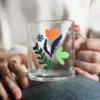 Manos de mujer sujetando sobre su pierna una taza de cristal personalizada con logo de colores