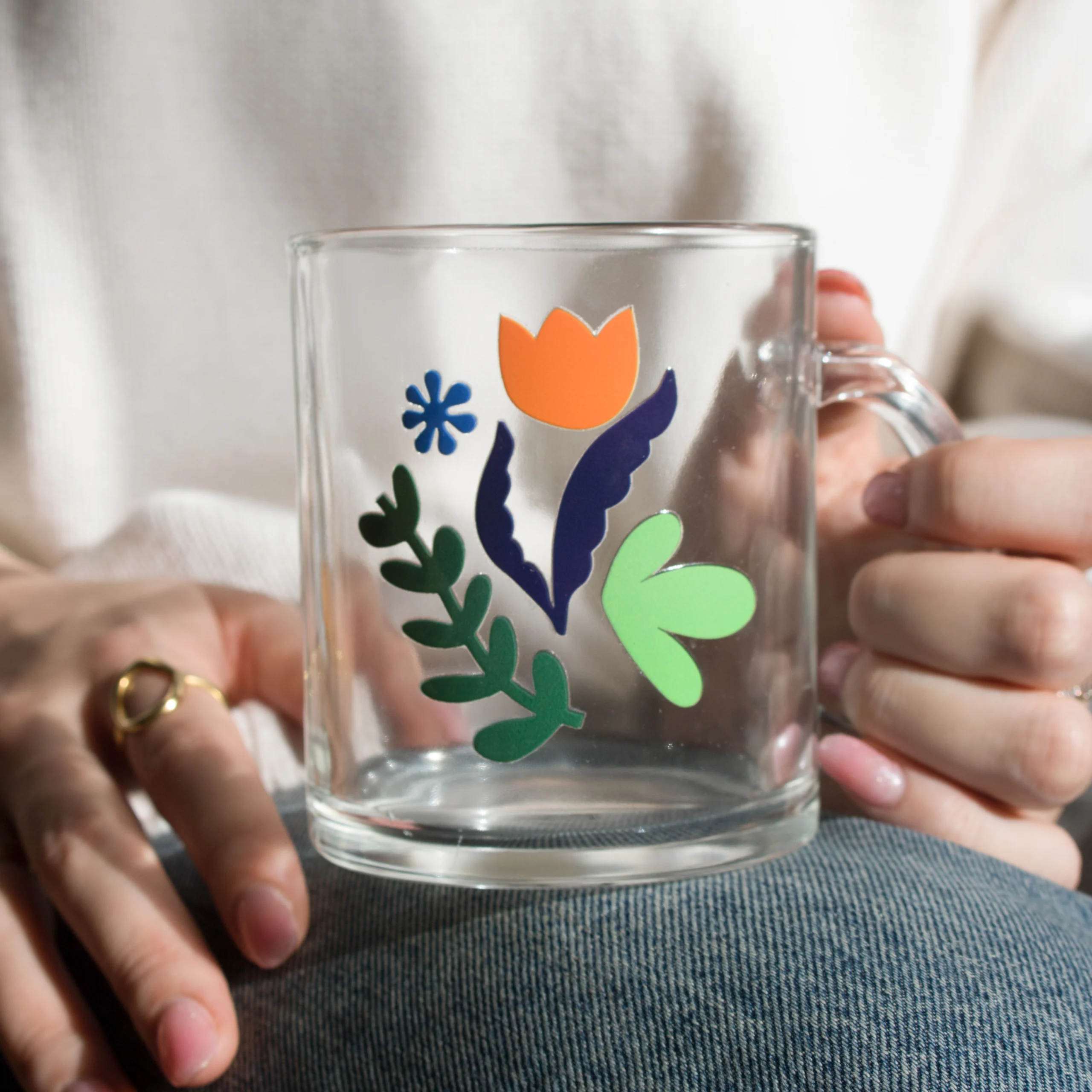Manos de mujer sujetando sobre su pierna una taza de cristal personalizada con logo de colores