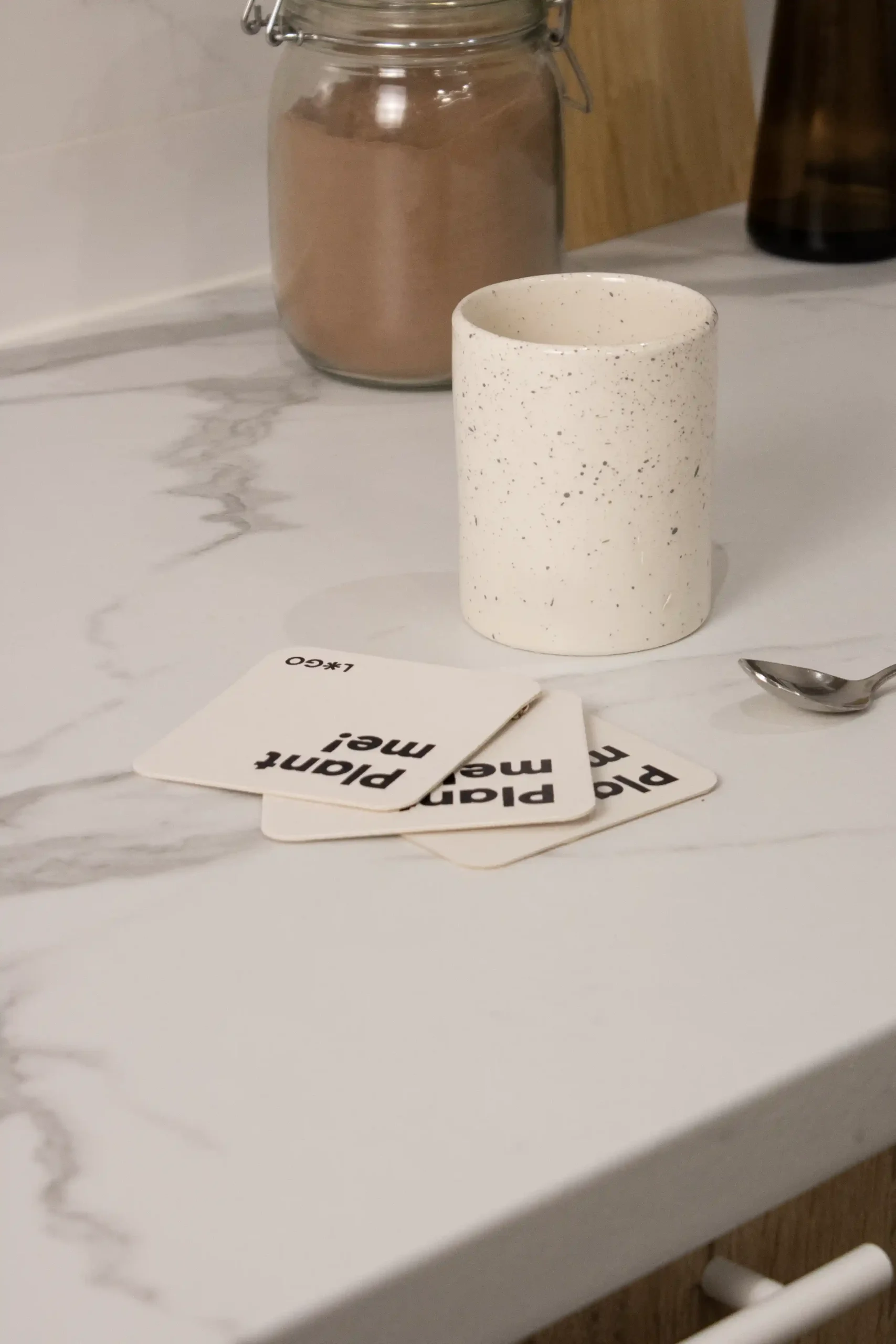 3 posavasos de cartón personalizados junto a una taza blanca y una cucharilla sobre la superficie de una cocina