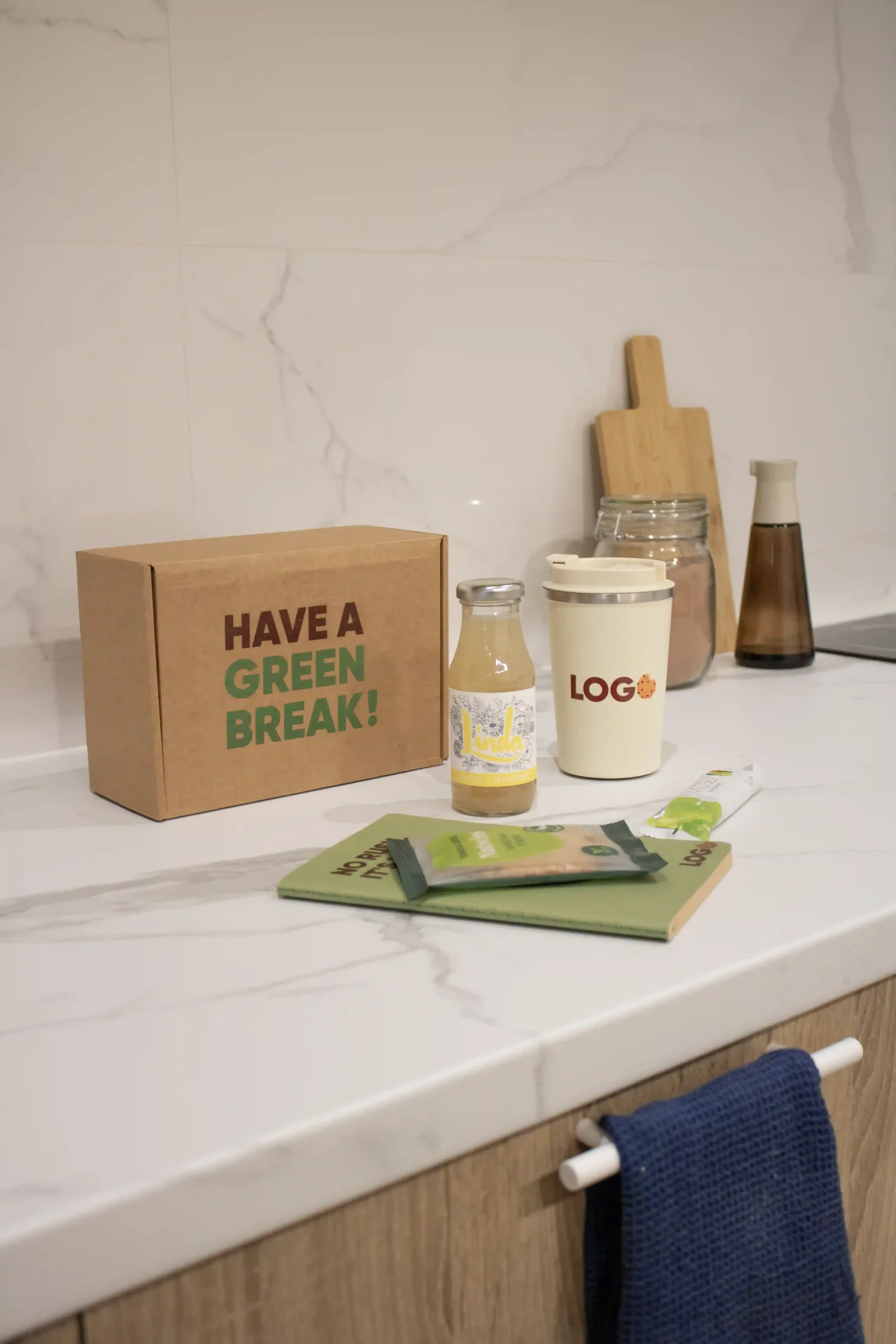 Un cuaderno reciclado verde, una botella con limonada, una taza de café take away, una galleta, una barrita de muesli y una caja de cartón personalizados y colocados sobre una superficie de cocina