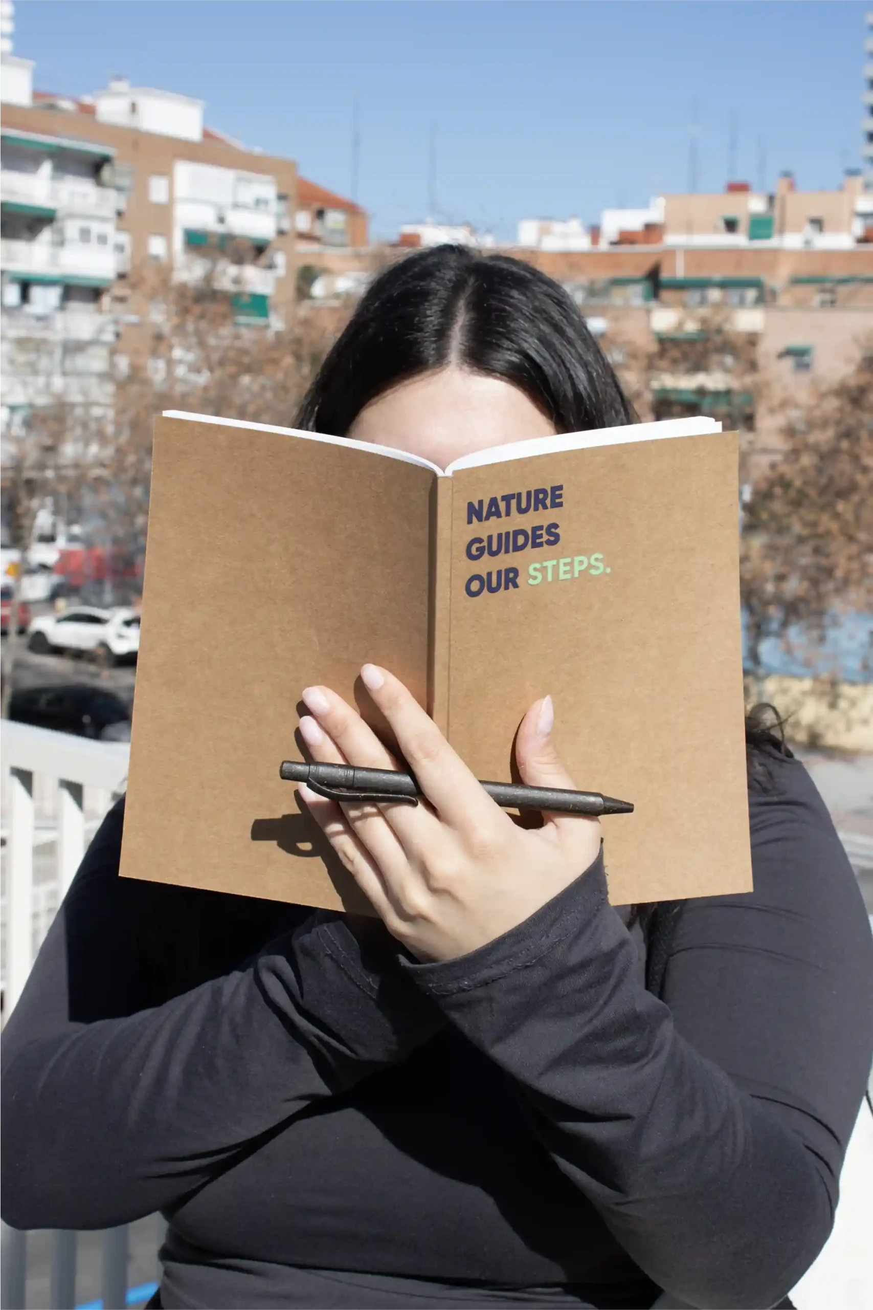 Chica sujetando un cuaderno ecológico kraft personalizado con el texto 'Nature guides our steps' y un bolígrafo marrón plantable