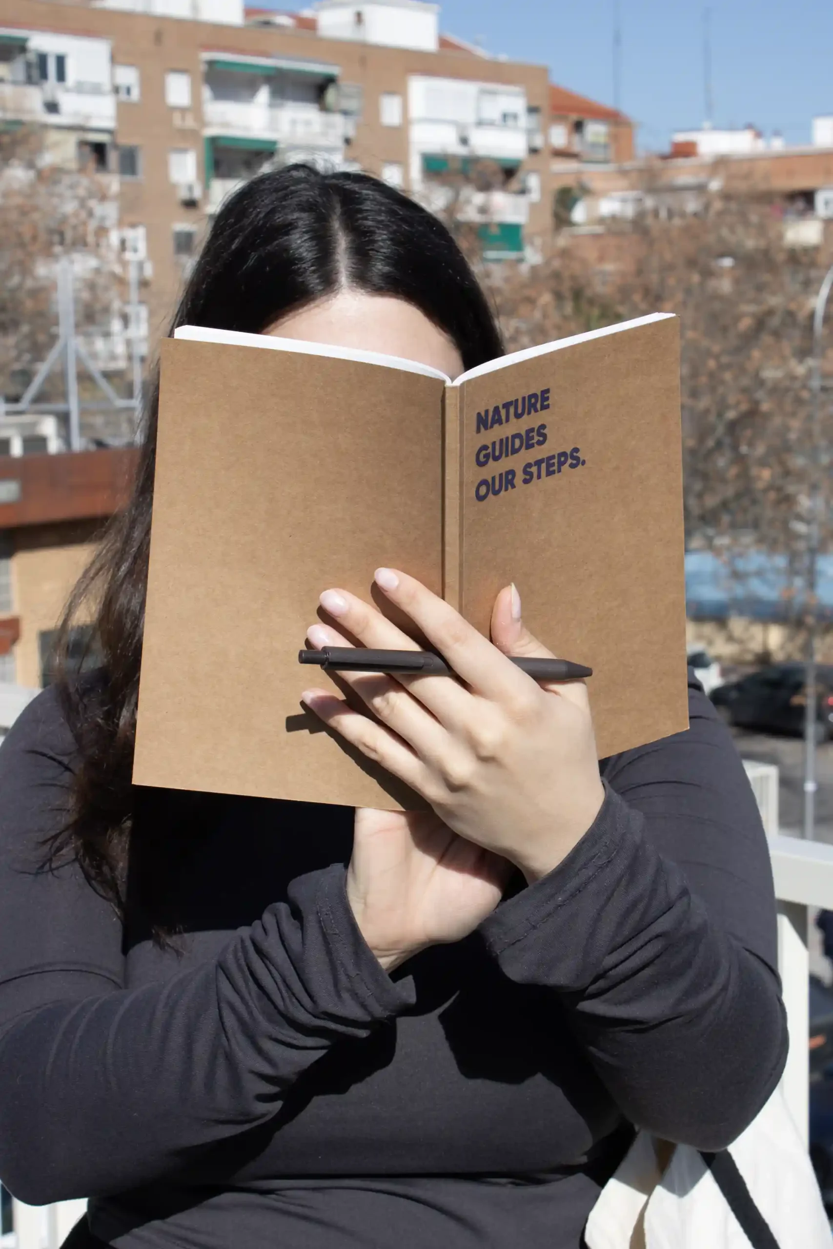 Chica sujetando un cuaderno plantable kraft personalizado con el texto 'Nature guides our steps' y un bolígrafo marrón plantable