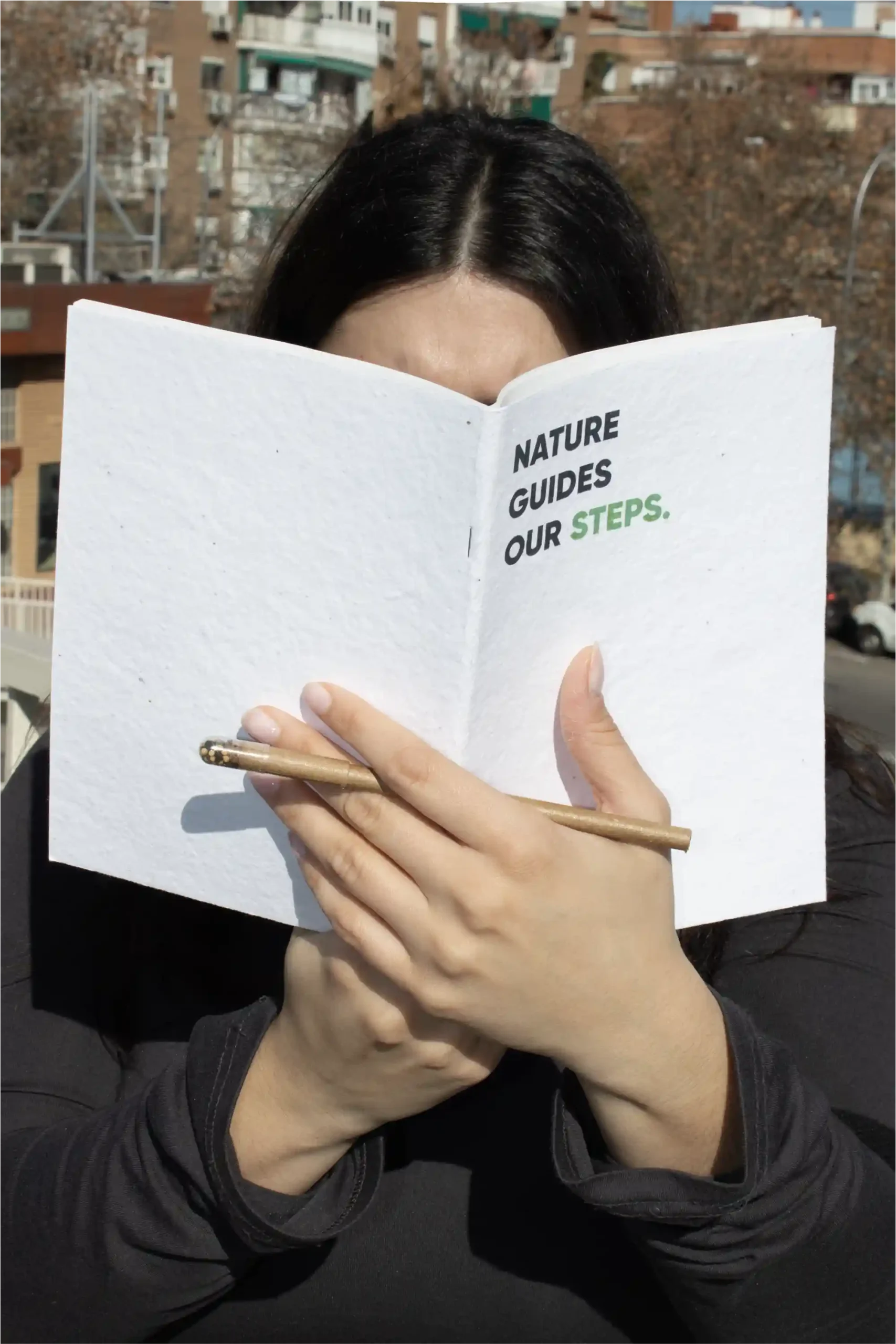 Chica sujetando un cuaderno plantable blanco personalizado con el texto 'Nature guides our steps' y un bolígrafo marrón plantable