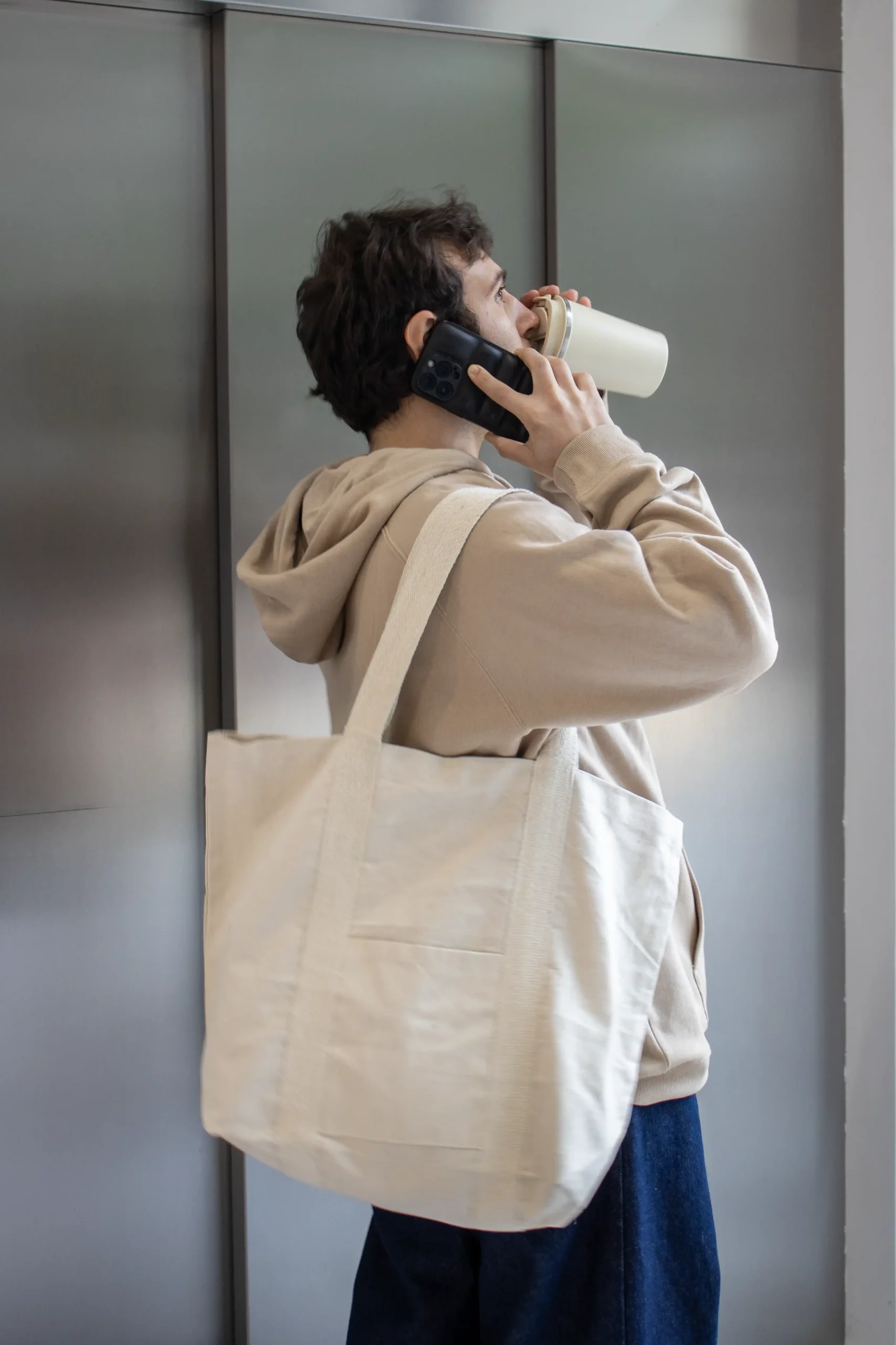 Chico moreno bebiendo de un termo y hablando por teléfono, llevando jeans y una sudadera beige con capucha personalizada