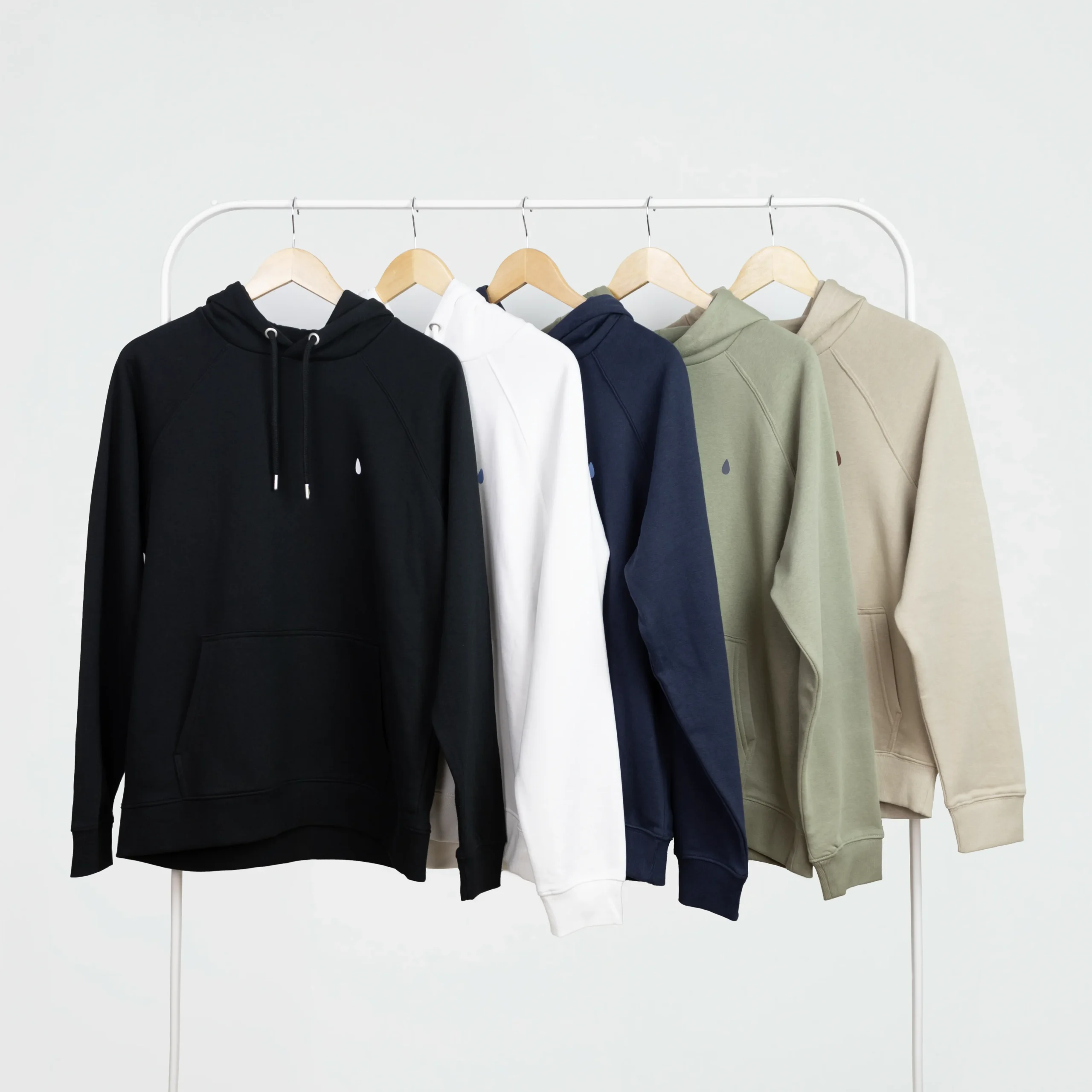 Cinco sudaderas con capucha personalizadas de diferentes colores, colgadas en perchas en un burro de ropa blanco