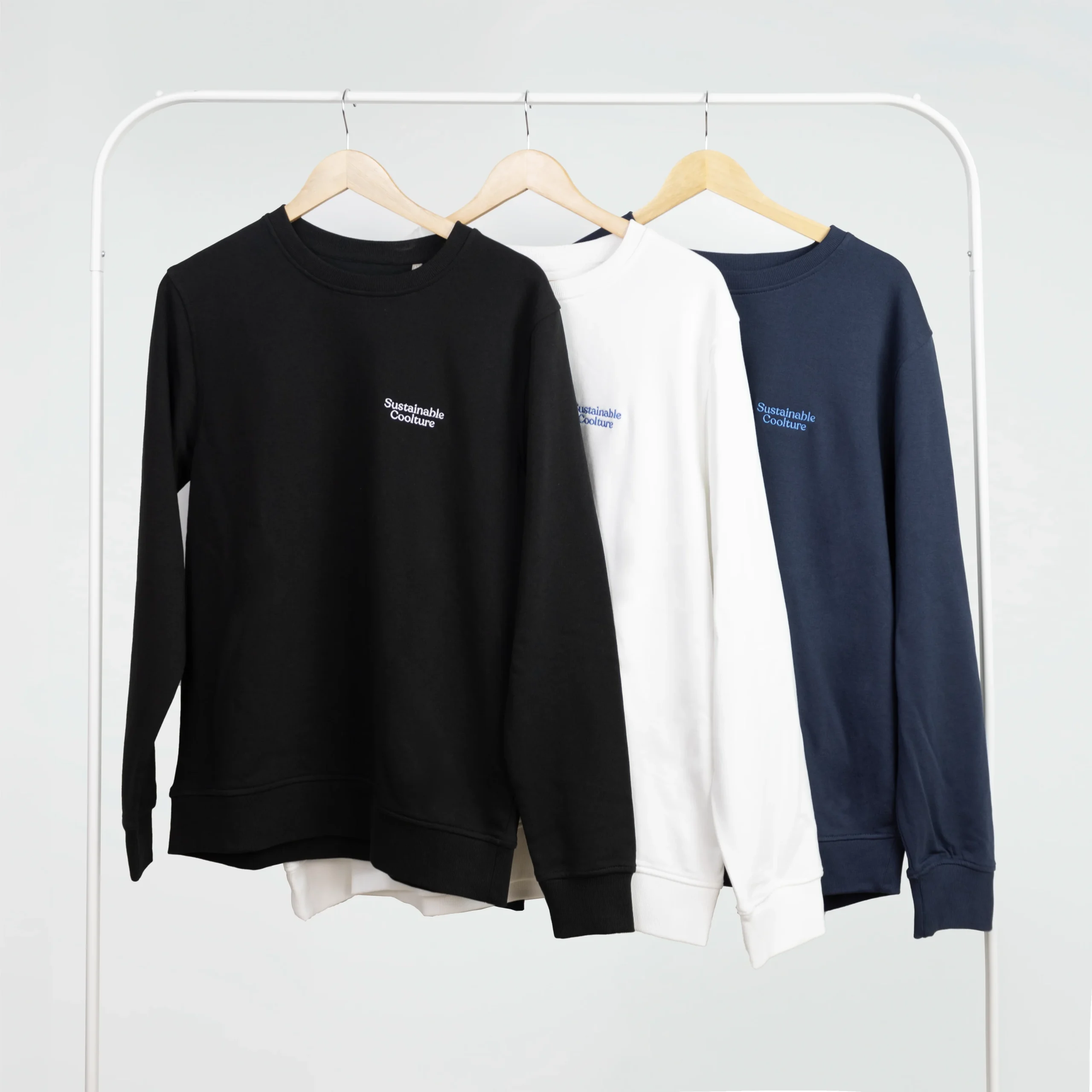 Tres sudaderas personalizadas de diferentes colores colgadas en perchas en un burro de ropa blanco