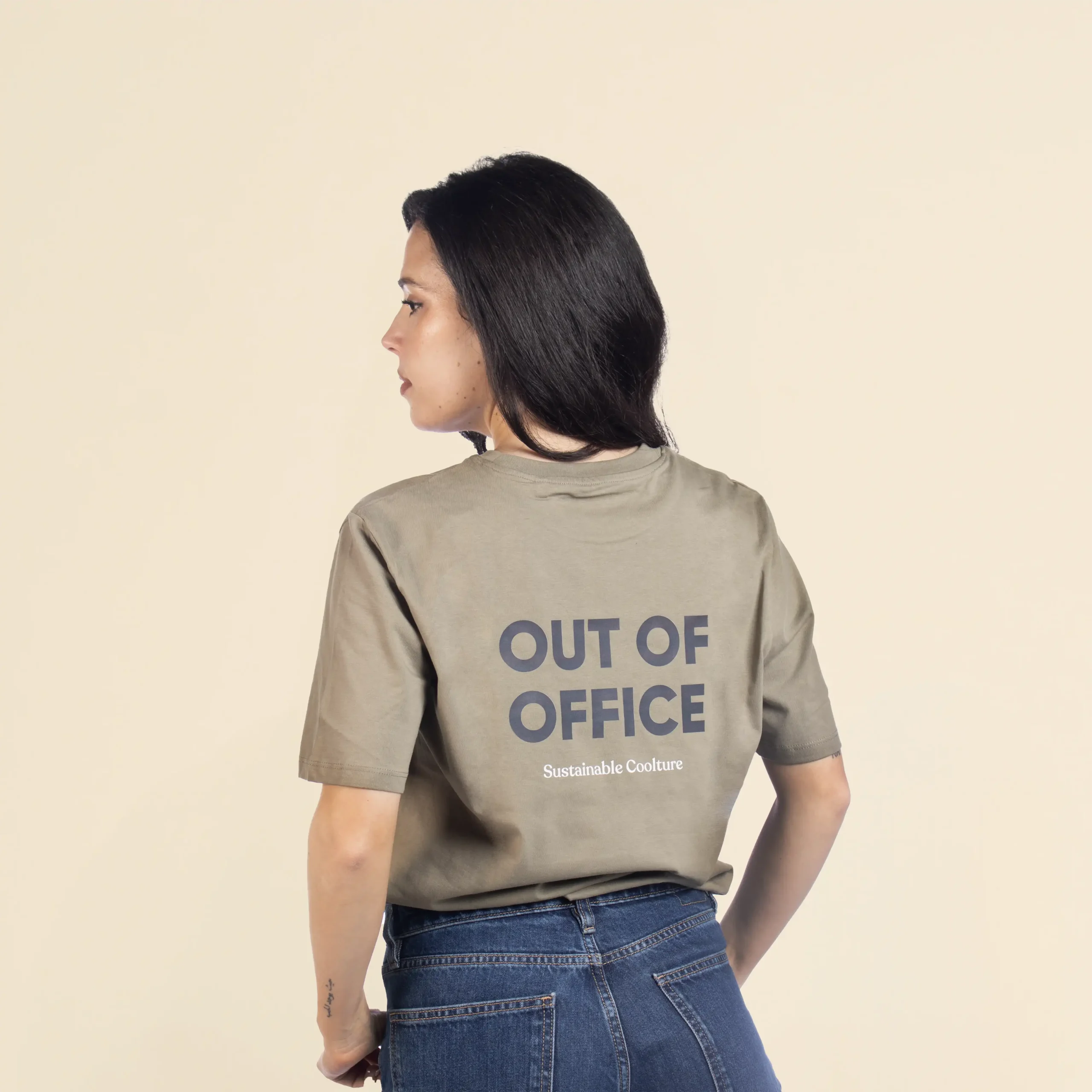 Chica morena de espaldas llevando jeans y una camiseta verde personalizada con el texto 'out of office'