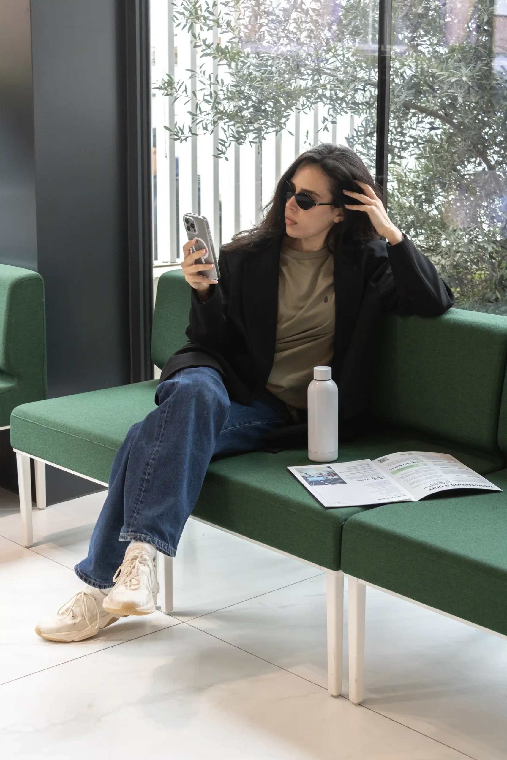 Chica morena con gafas de sol, sentada en un sofa mirando el móvil, llevando jeans, una camiseta verde personalizada y una americana negra