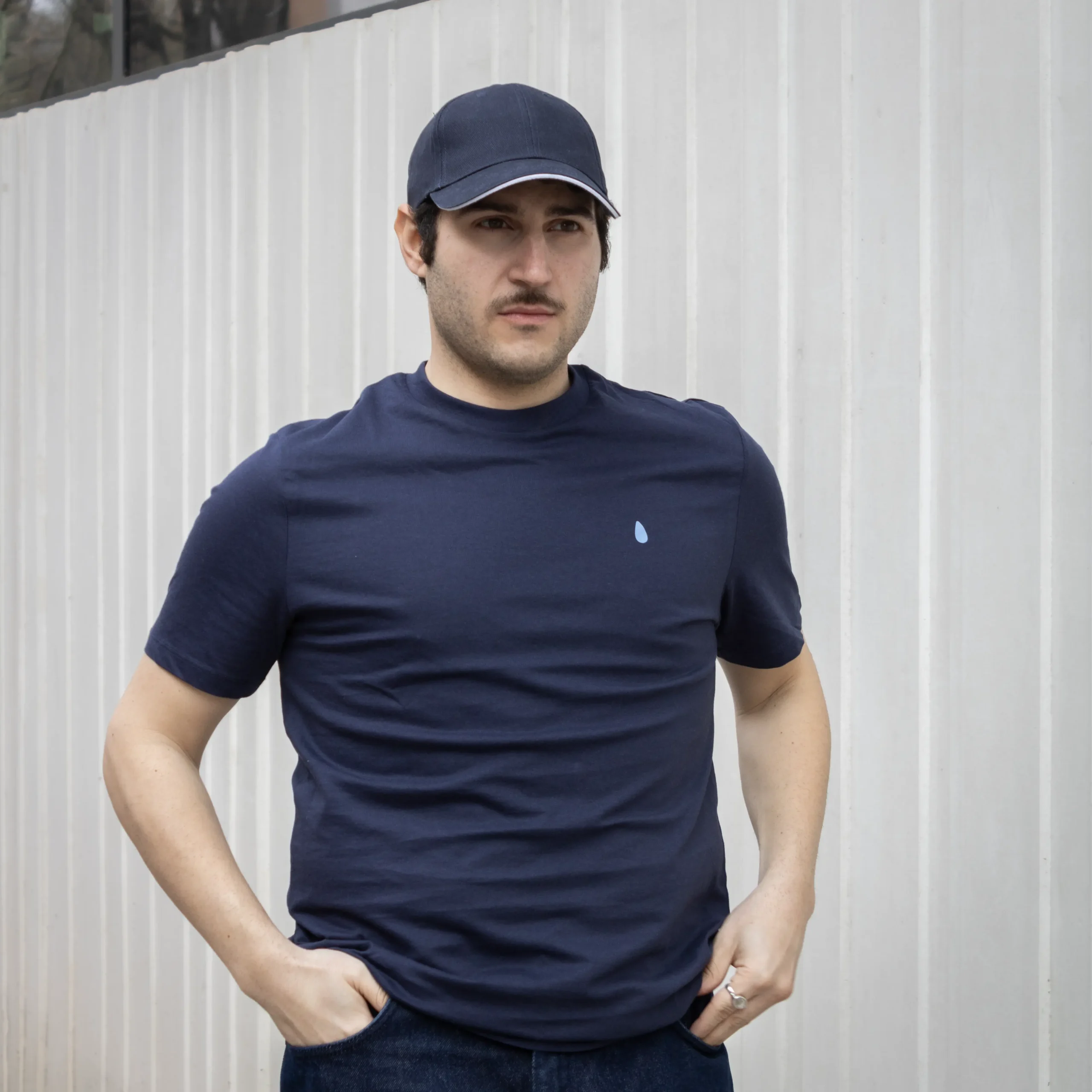 Chico moreno con gorra posando en la calle, llevando jeans y una camiseta azul marino personalizada