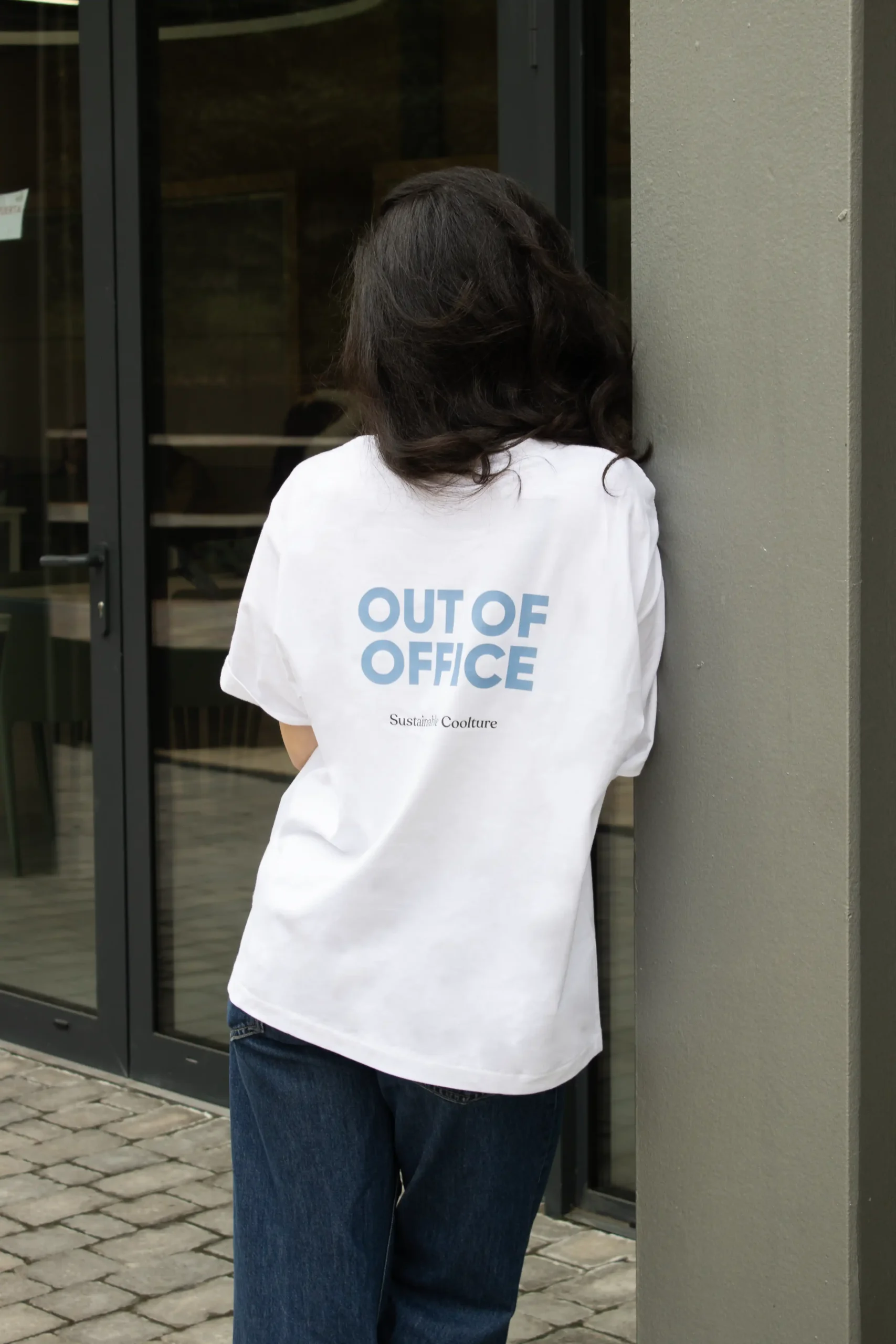 Chica morena de espaldas llevando jeans y una camiseta oversize blanca personalizada con el texto "out of office"