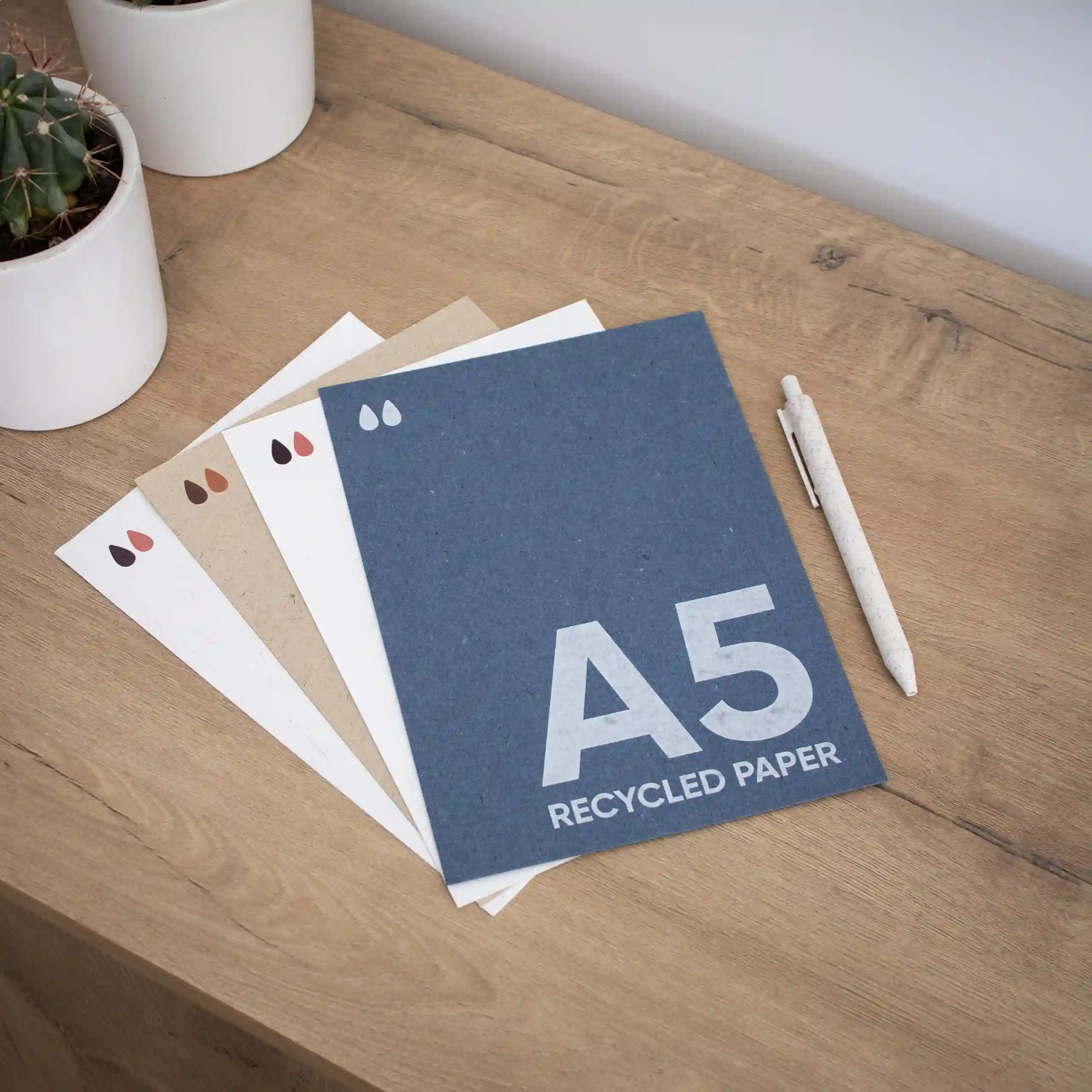 Cuatro tarjetas A5 ecológicas junto a un bolígrafo sobre una superficie de madera