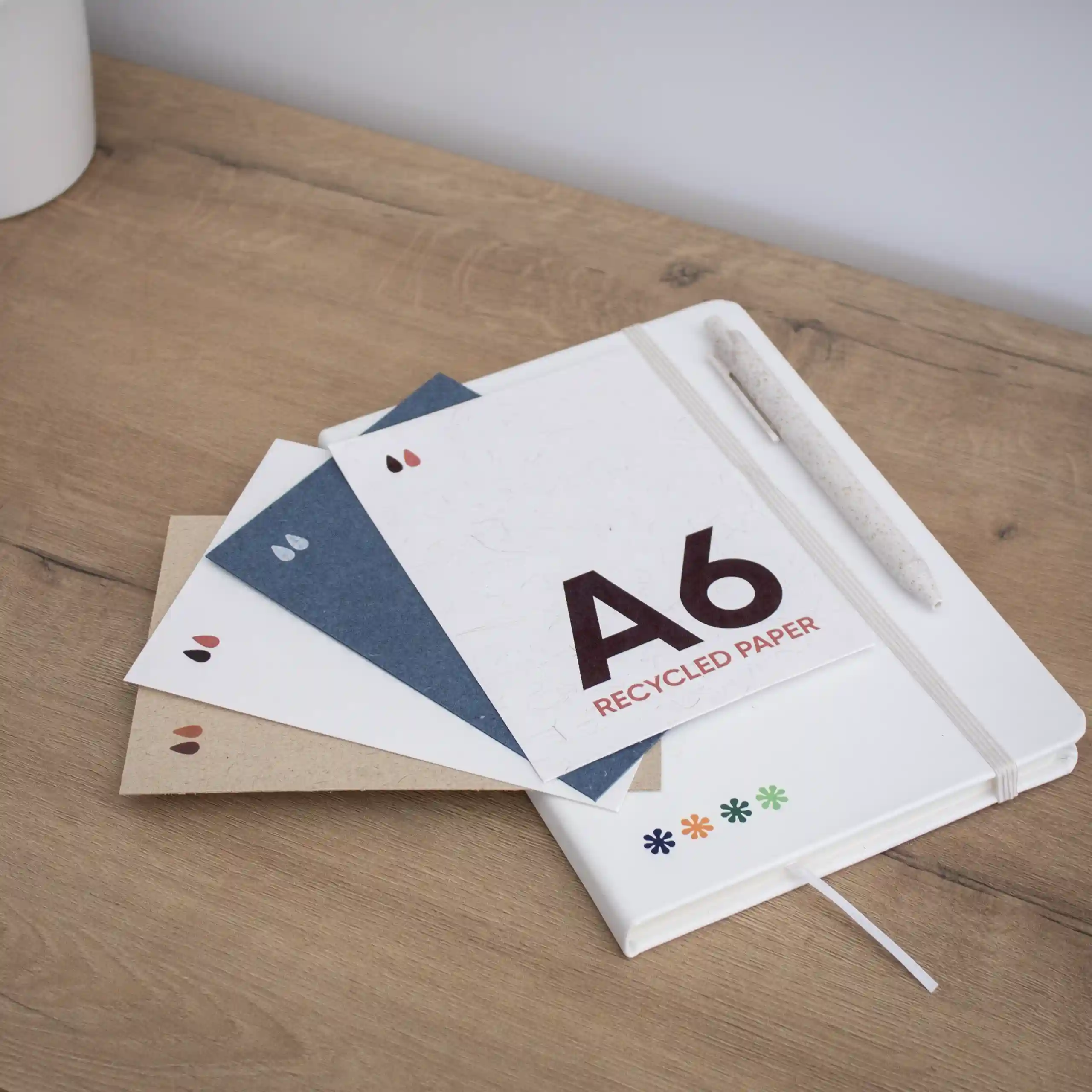 cuatro tarjetas A6 impresas en papeles ecológicos junto a un cuaderno blanco y un bolígrafo, sobre una superficie de madera