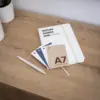 Cuatro tarjetas A7 impresas en papeles ecológicos junto a un cuaderno blanco y un bolígrafo, sobre una superficie de madera