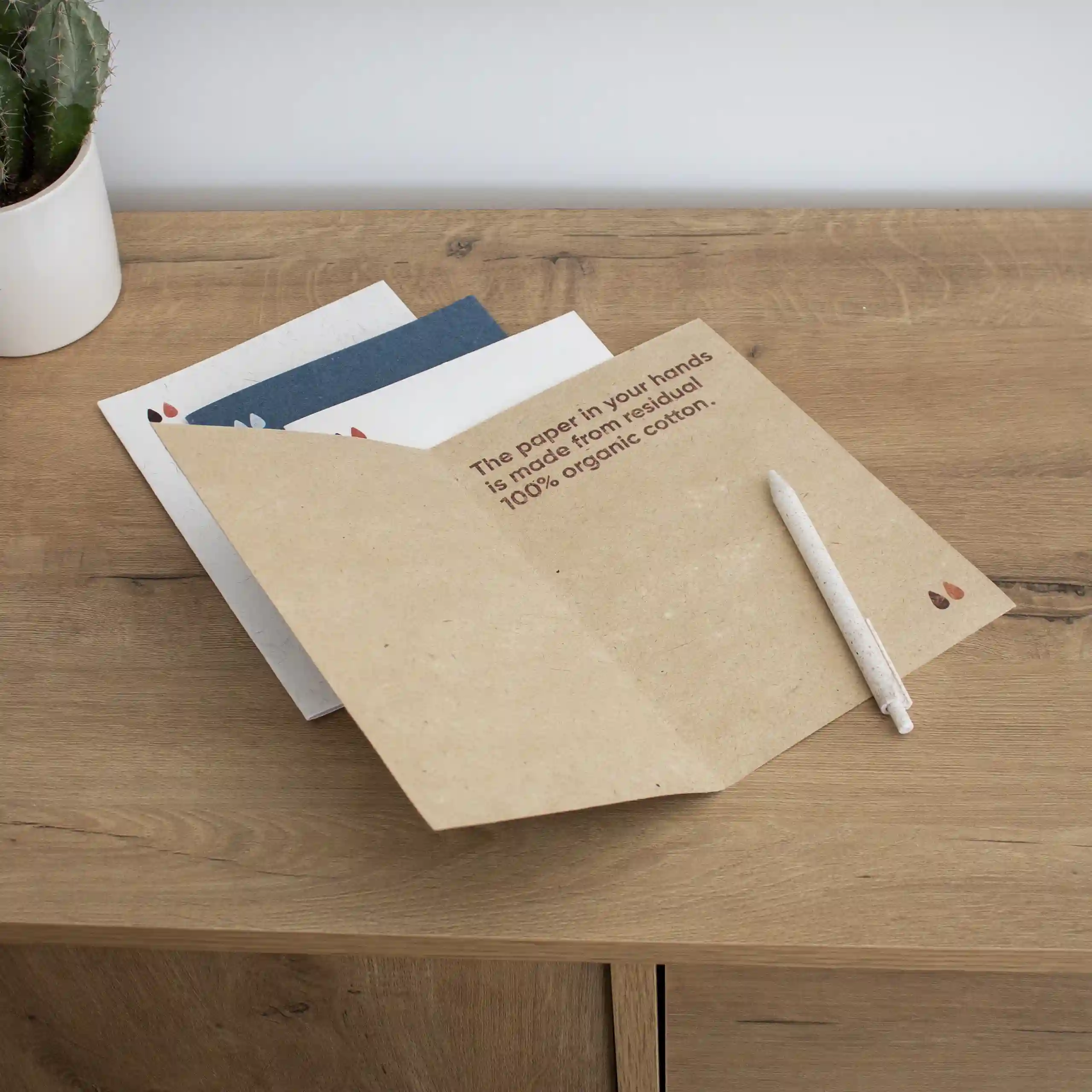 Cuatro dípticos A5 impresos en papel ecológico junto con un bolígrafo sobre una superficie de madera