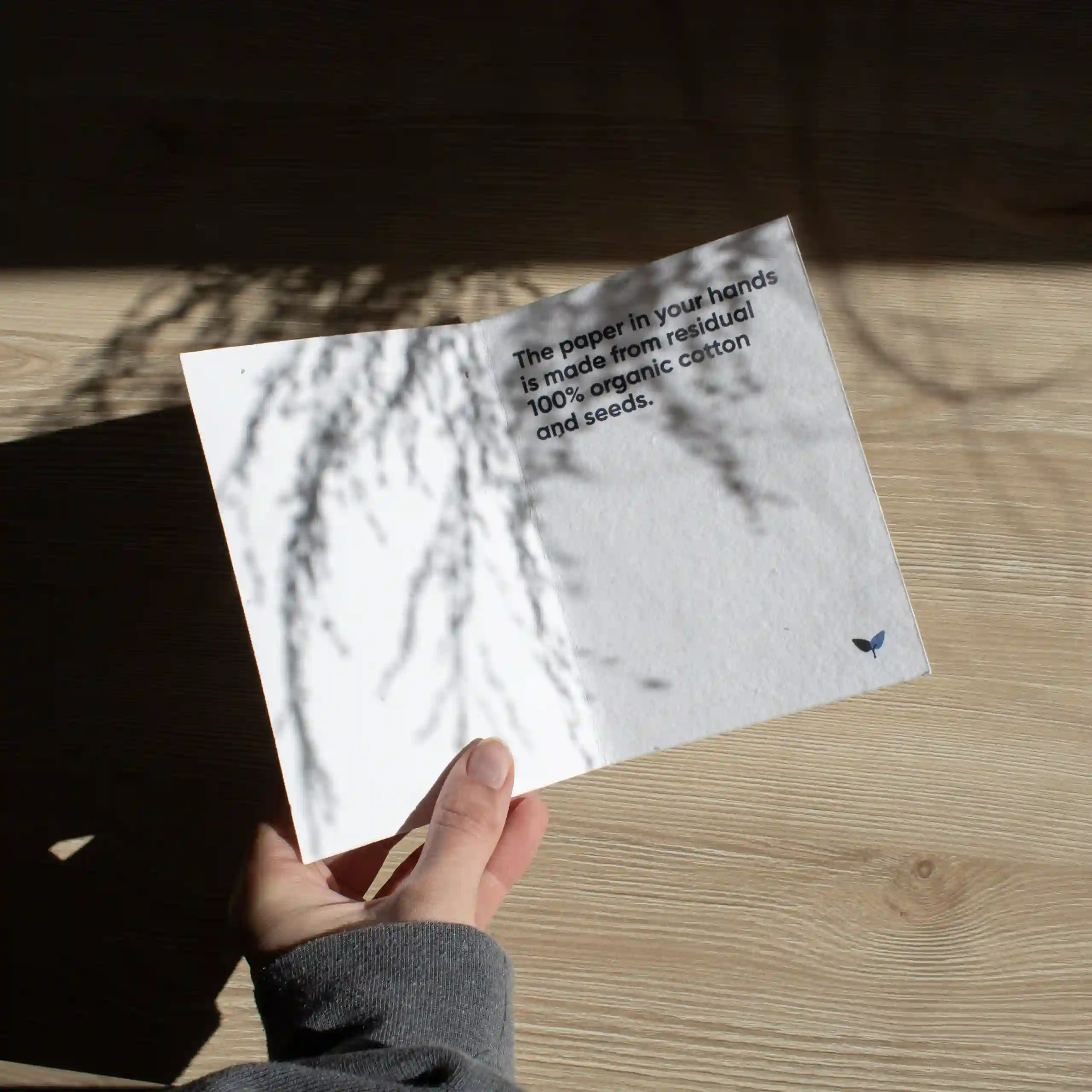 Díptico A6 plantable Mano sujetando un díptico A6 abierto impreso en papel plantable sobre una superficie de madera con sombras