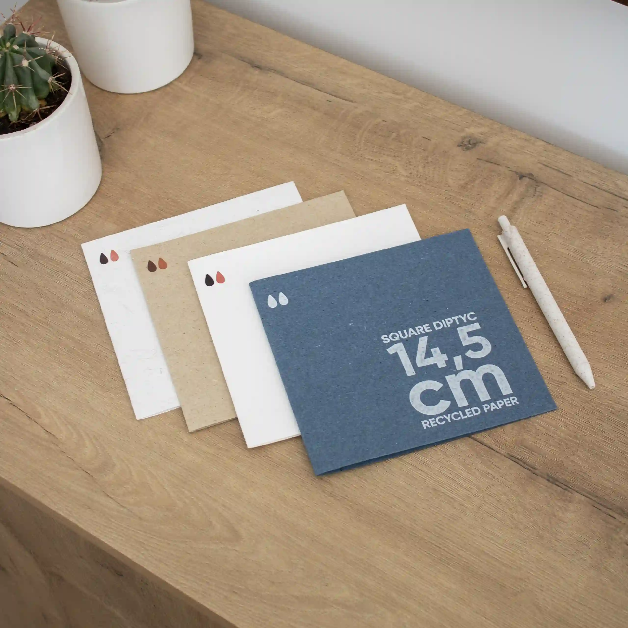 Cuatro dípticos cuadrados impresos en papel ecológico junto a un bolígrafo sobre un superficie de madera