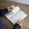 Dos manos colocando un marcapágina de papel plantable dentro de un cuaderno sobre una superficie de madera