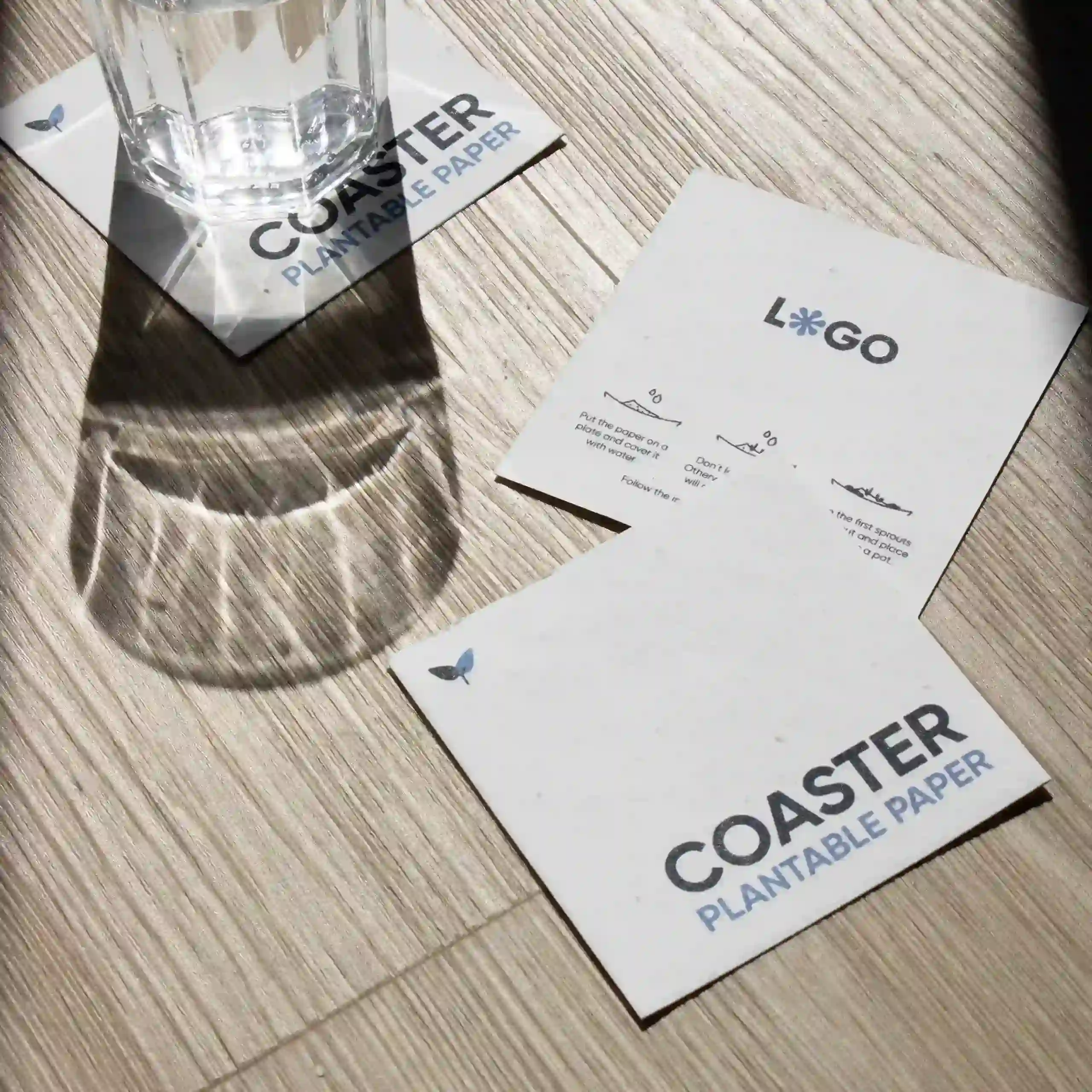 Posavasos de papel plantable Tres posavasos de papel plantable personalizados y un vaso de cristal sobre una superficie de madera al sol